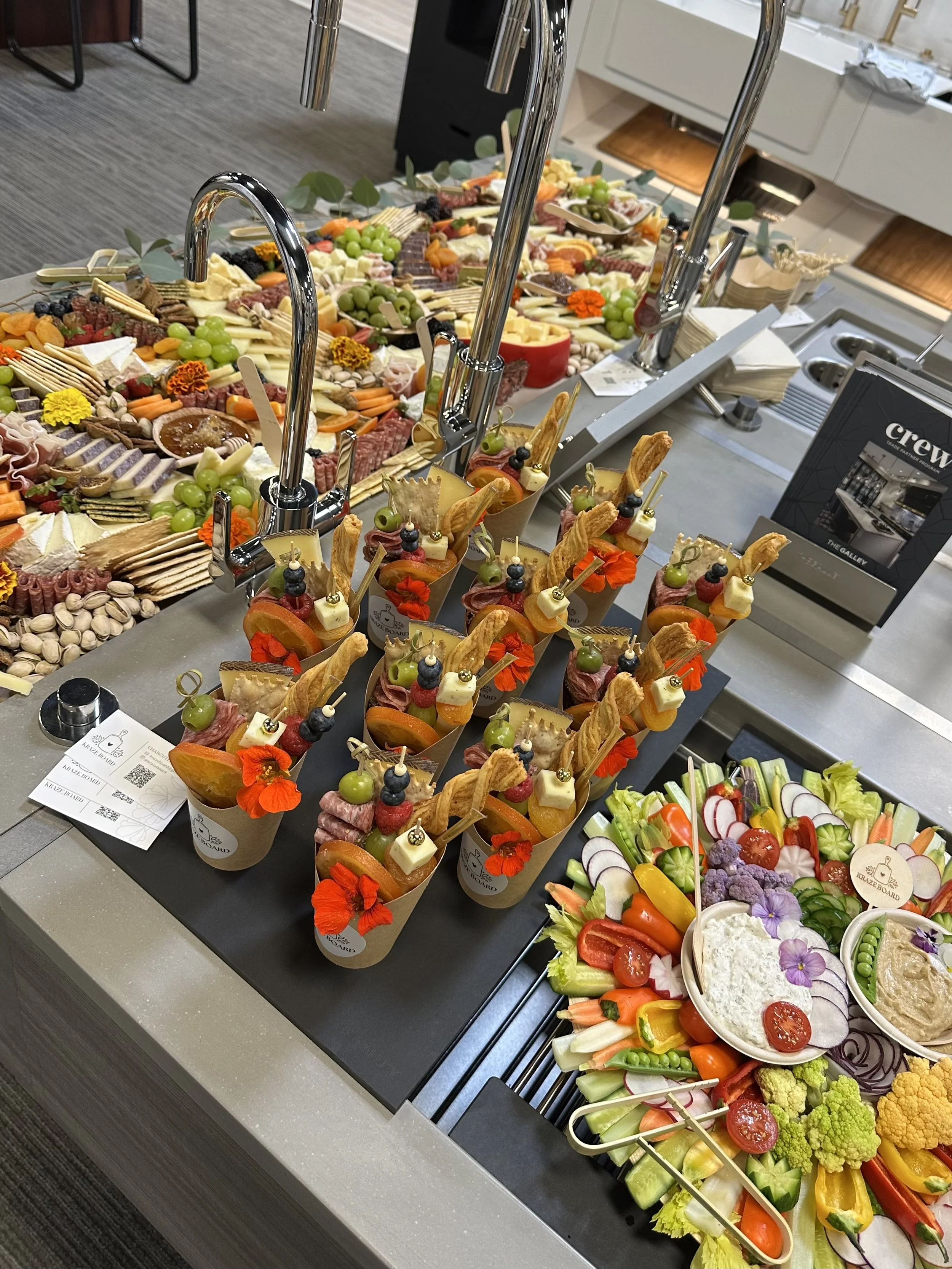 A buffet table with an assortment of appetizers, including vegetables, cheeses, meats, and fruit, with small cups containing finger foods decorated with edible flowers and garnishes.