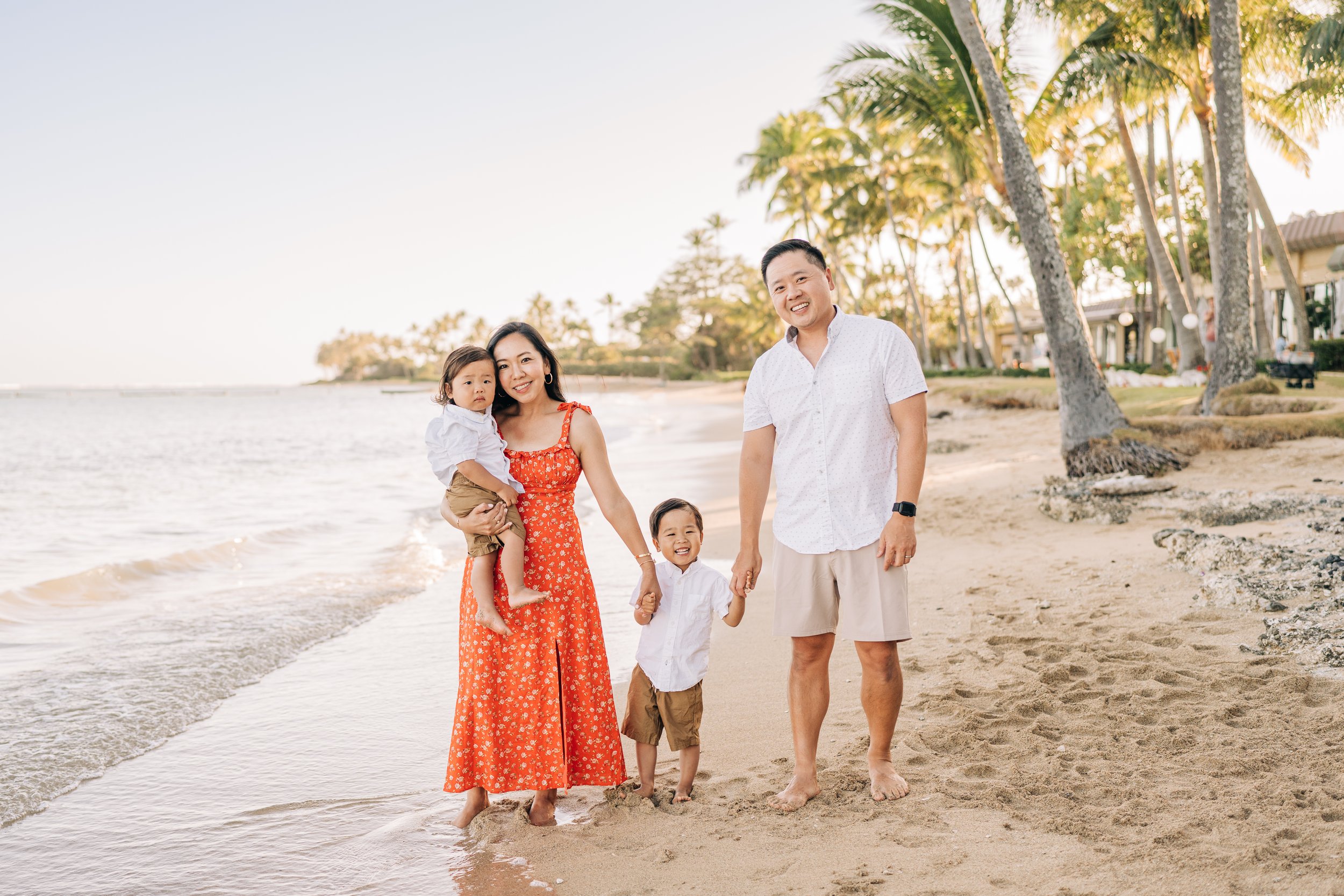 A family of four enjoying a walk on the beach during sunset, with palm trees and beach houses in the background.