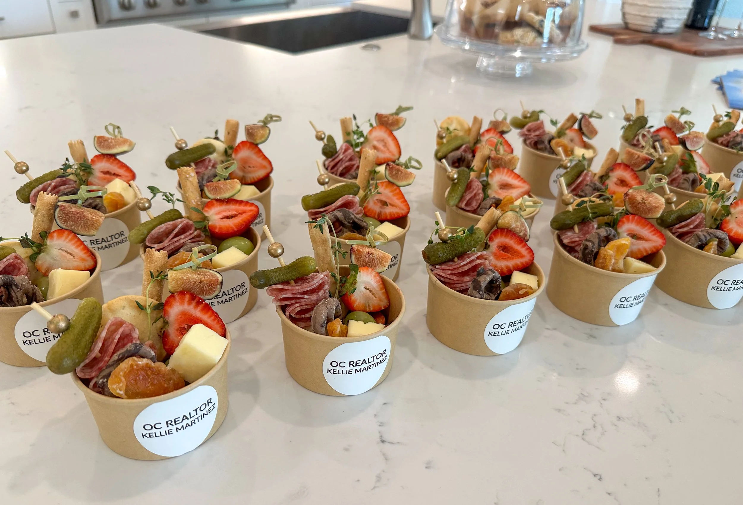 Multiple small cups with assorted cheeses, strawberries, pickles, and cured meats, each with a decorative toothpick, on a white countertop.