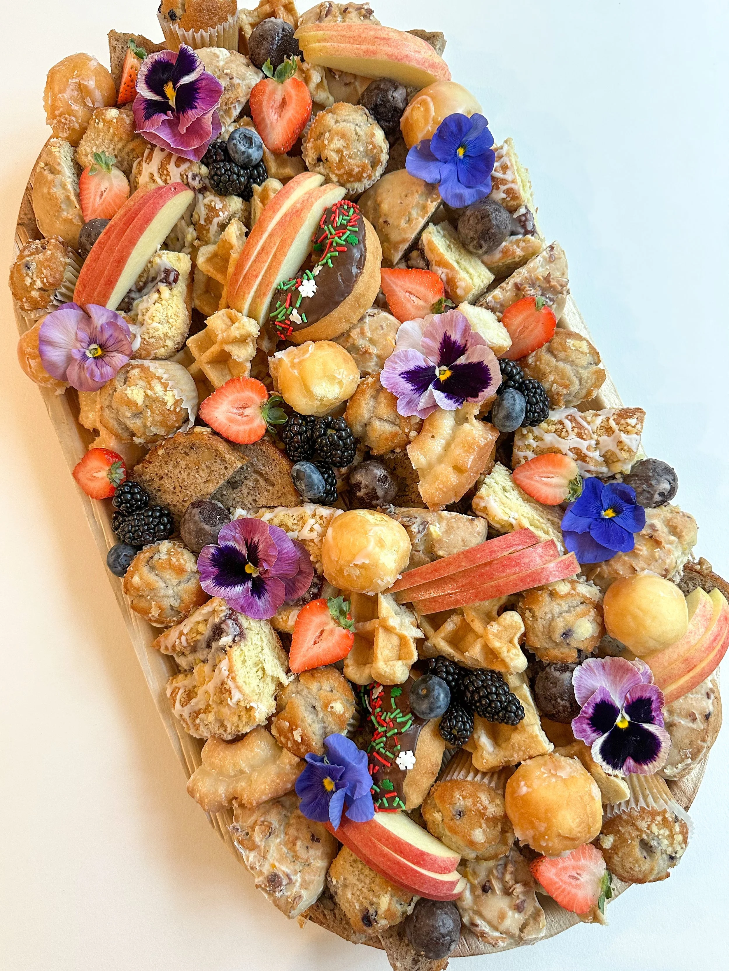 A large fruit and dessert platter with various fruits, cookies, pastries, edible flowers, and a donut with sprinkles.