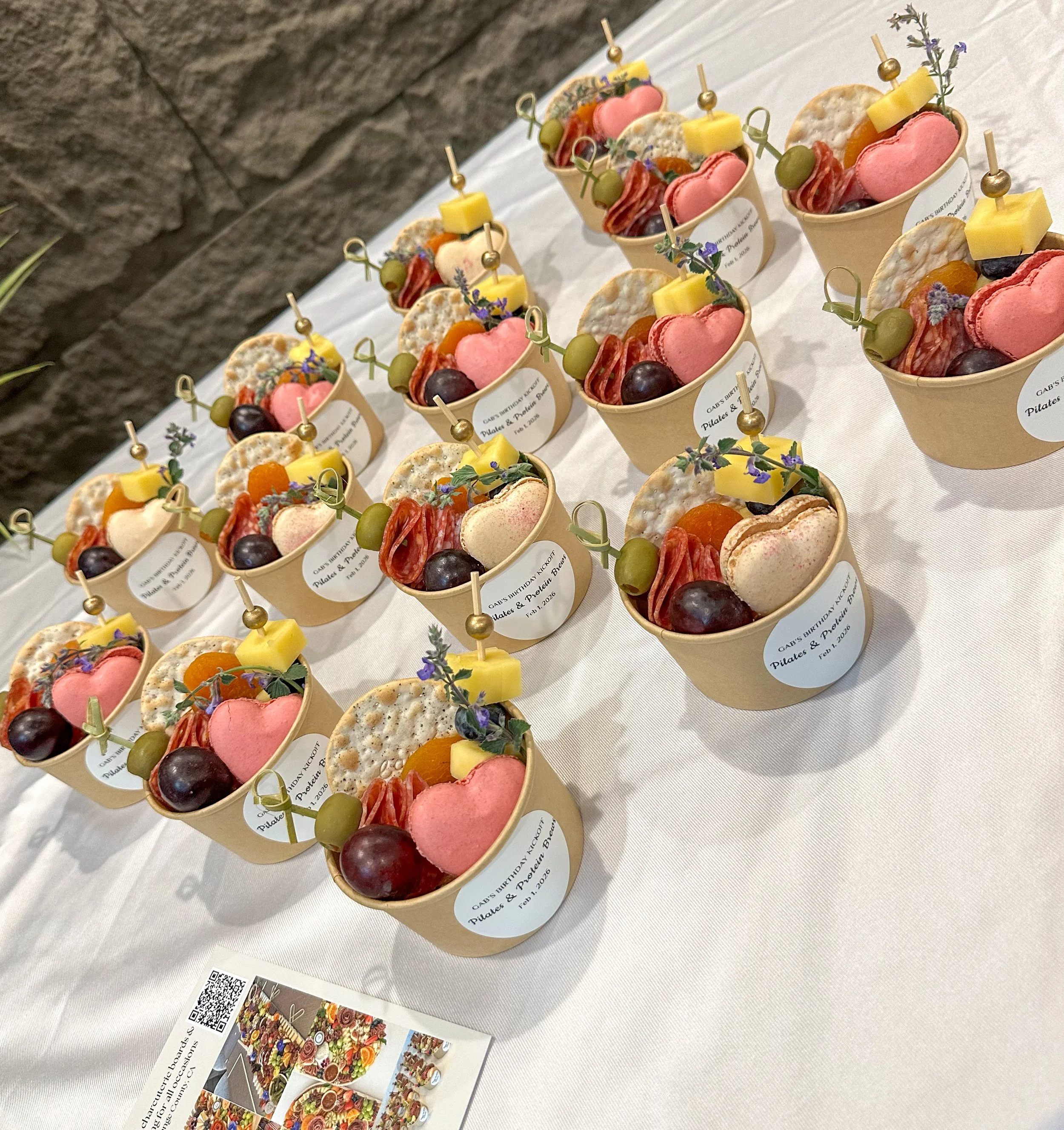Multiple small cups with assorted cheeses, strawberries, pickles, and cured meats, each with a decorative toothpick, on a white countertop.