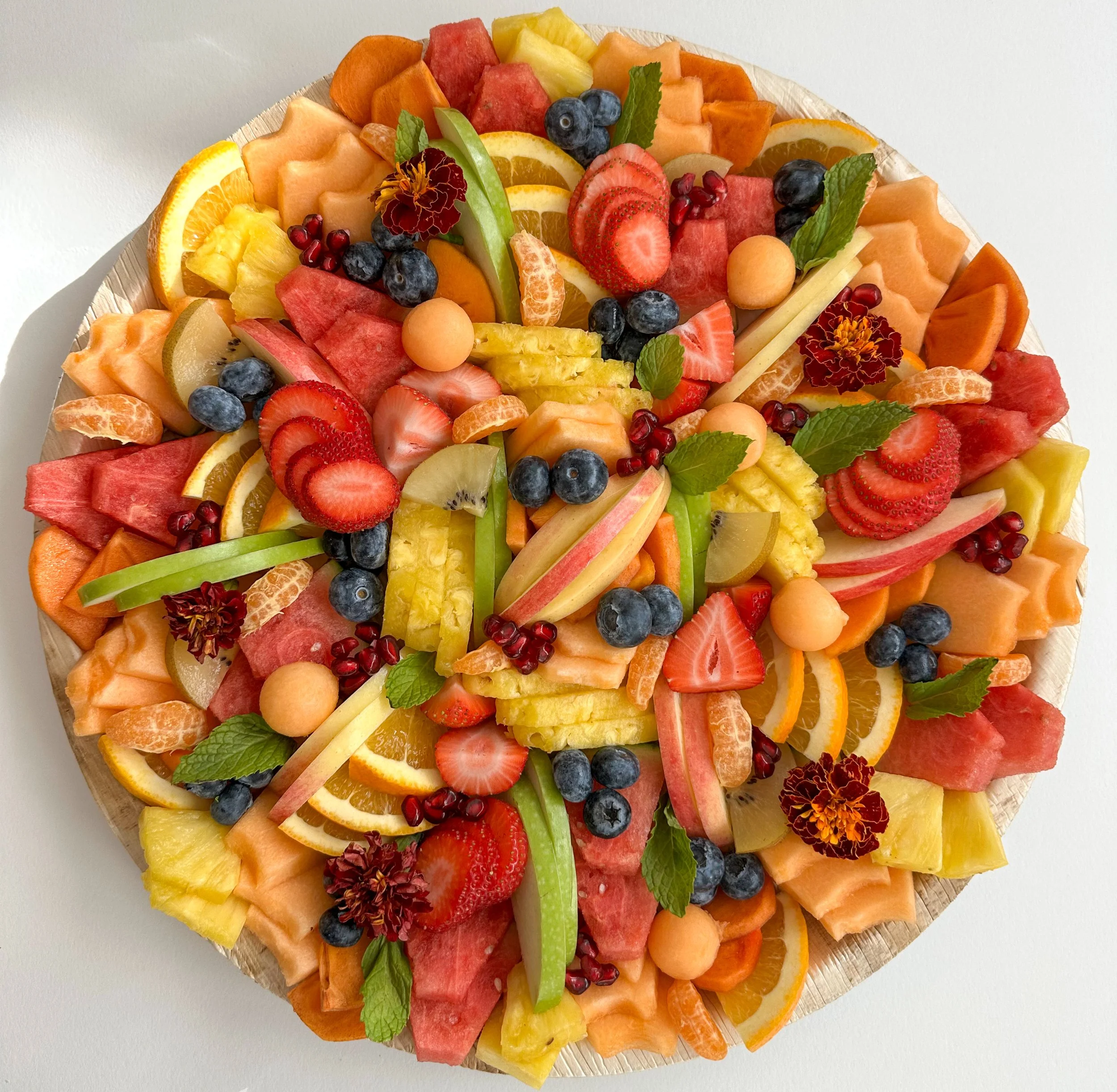 Colorful assorted sliced fresh fruits on a round wooden platter, including watermelon, strawberries, blueberries, pineapple, kiwi, cantaloupe, honeydew, orange slices, apple slices, pomegranate seeds, and garnished with mint leaves and edible flowers.