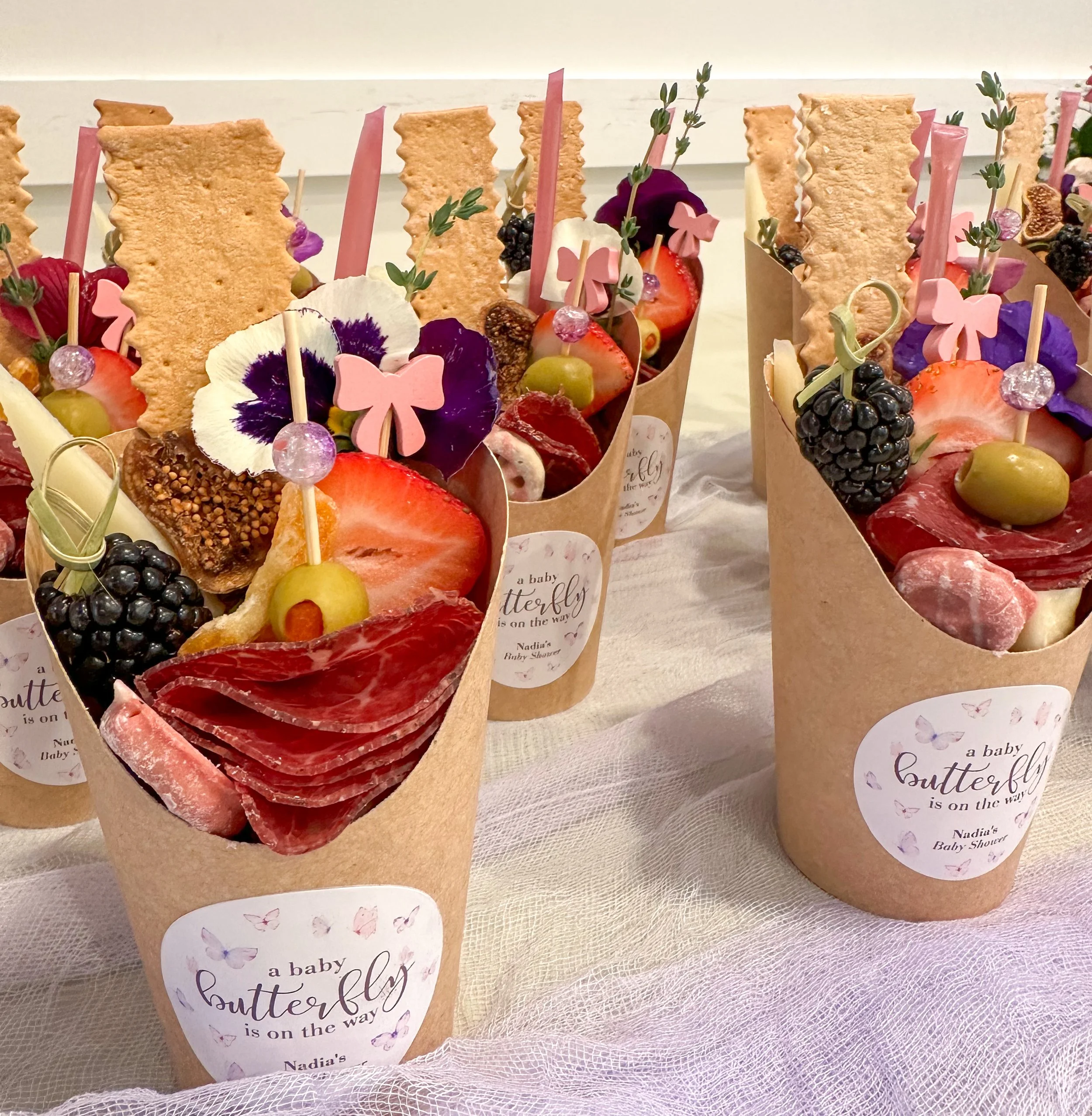 A platter of colorful fruit and meat appetizers garnished with edible flowers, cheese, and crackers, topped with skewers of grapes and berries.