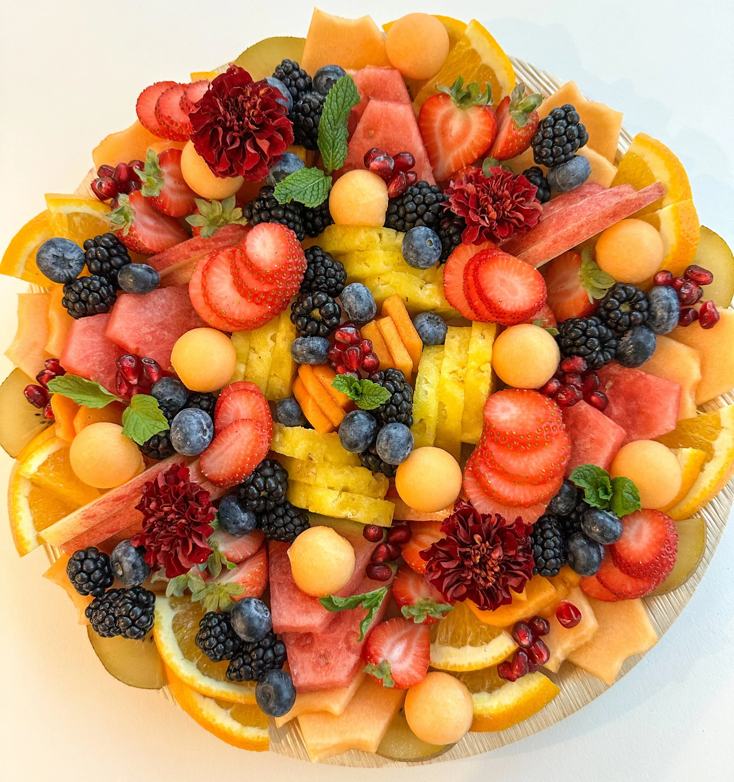 A colorful fruit platter featuring strawberries, blackberries, blueberries, watermelon, cantaloupe, honeydew, orange slices, pomegranate seeds, kiwi slices, and garnished with mint leaves.