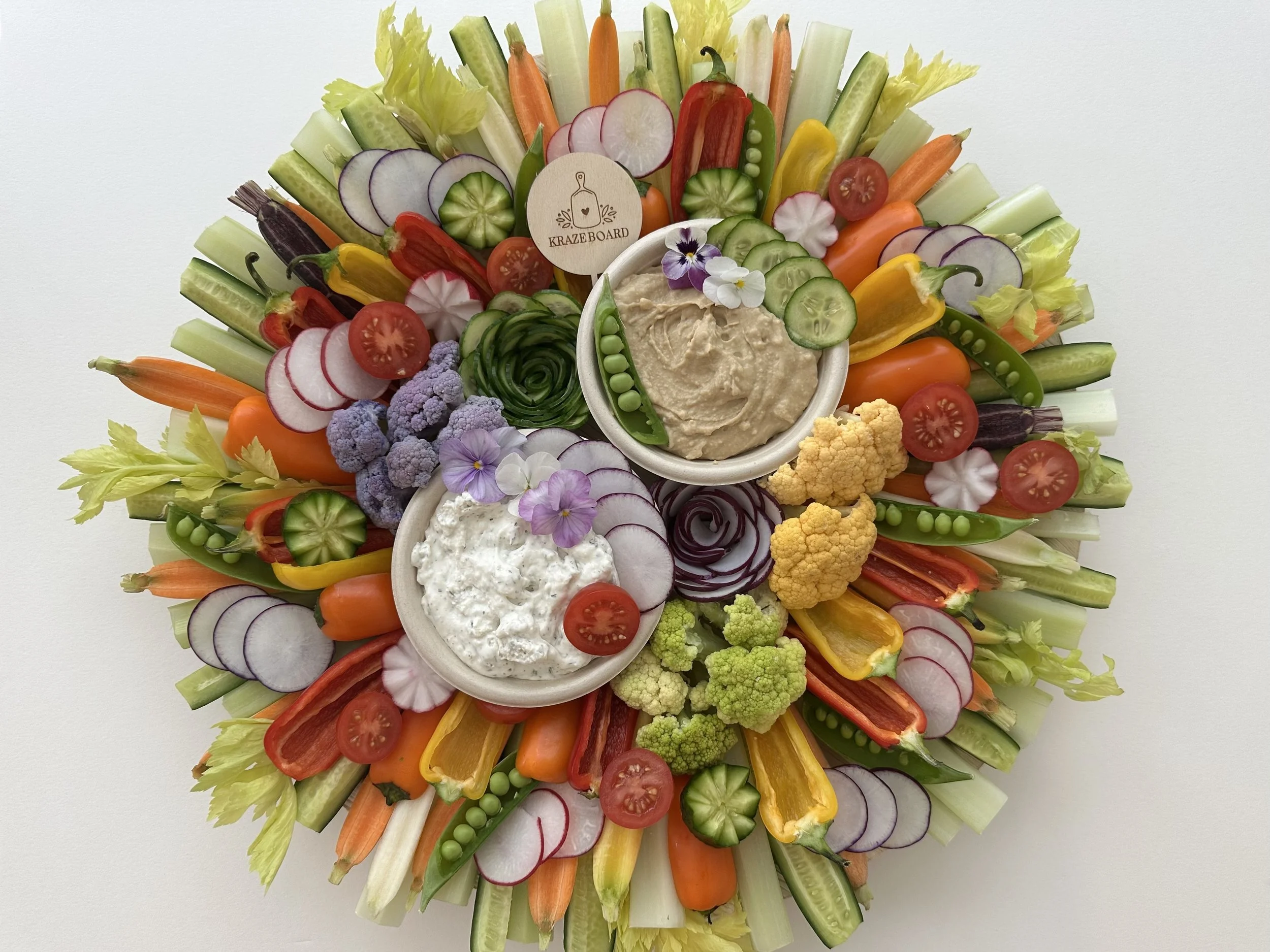 Colorful vegetable platter with cherry tomatoes, cucumber slices, radish slices, bell peppers, cauliflower, broccoli, and two bowls of creamy dips decorated with edible flowers.