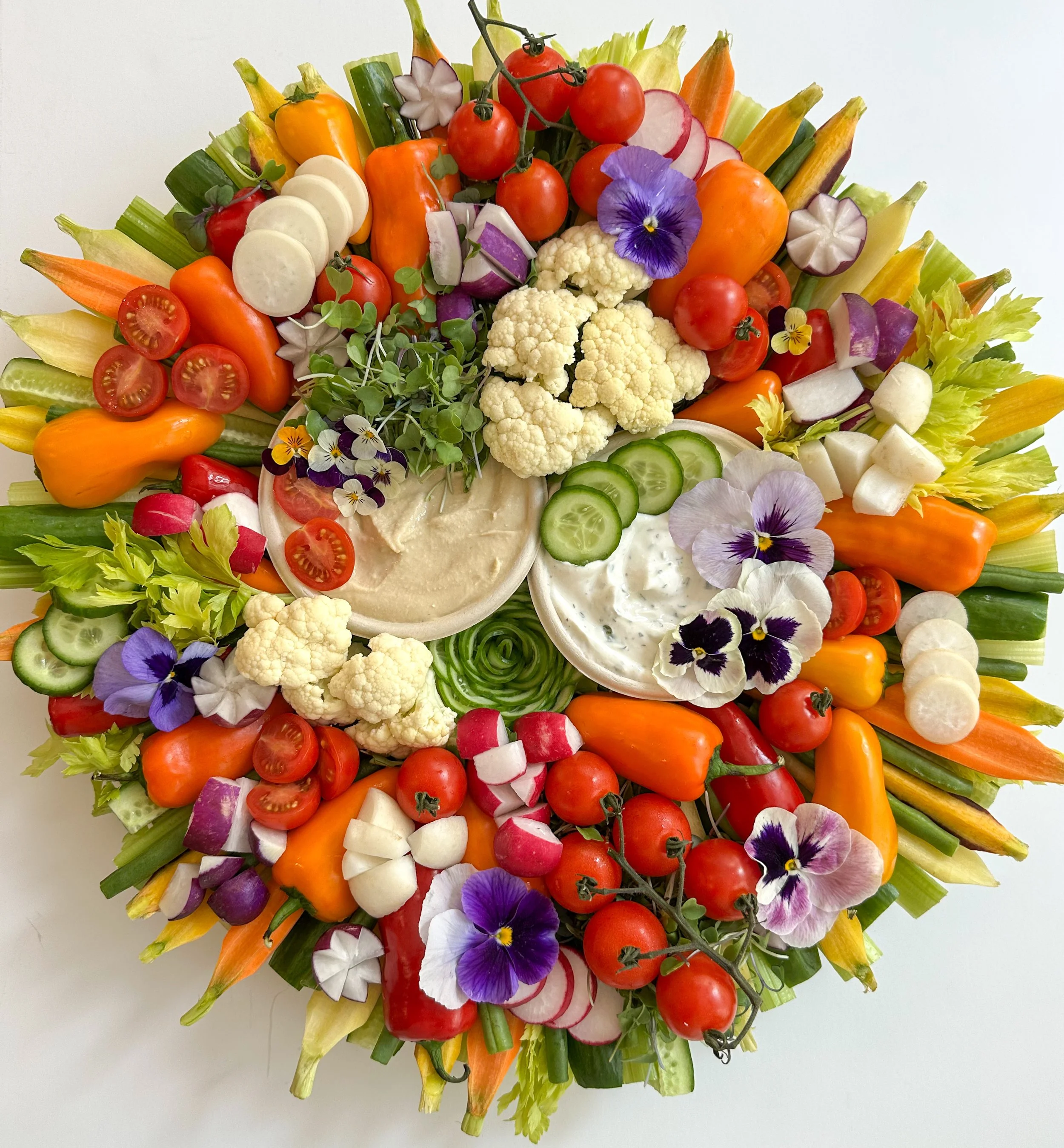 Colorful vegetable platter with cherry tomatoes, cucumber slices, radish slices, bell peppers, cauliflower, broccoli, and two bowls of creamy dips decorated with edible flowers.