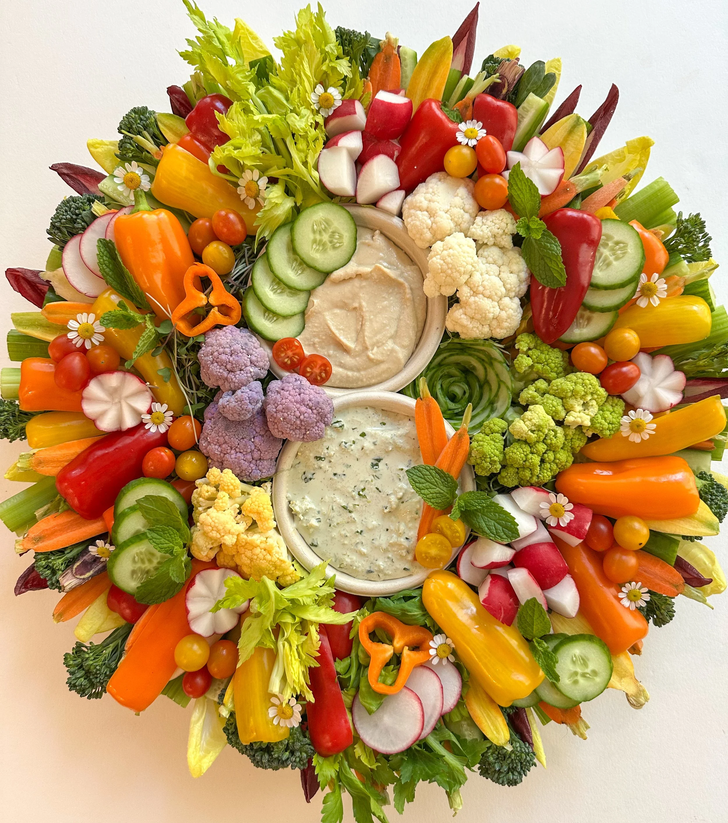 Colorful vegetable platter with cherry tomatoes, cucumber slices, radish slices, bell peppers, cauliflower, broccoli, and two bowls of creamy dips decorated with edible flowers.