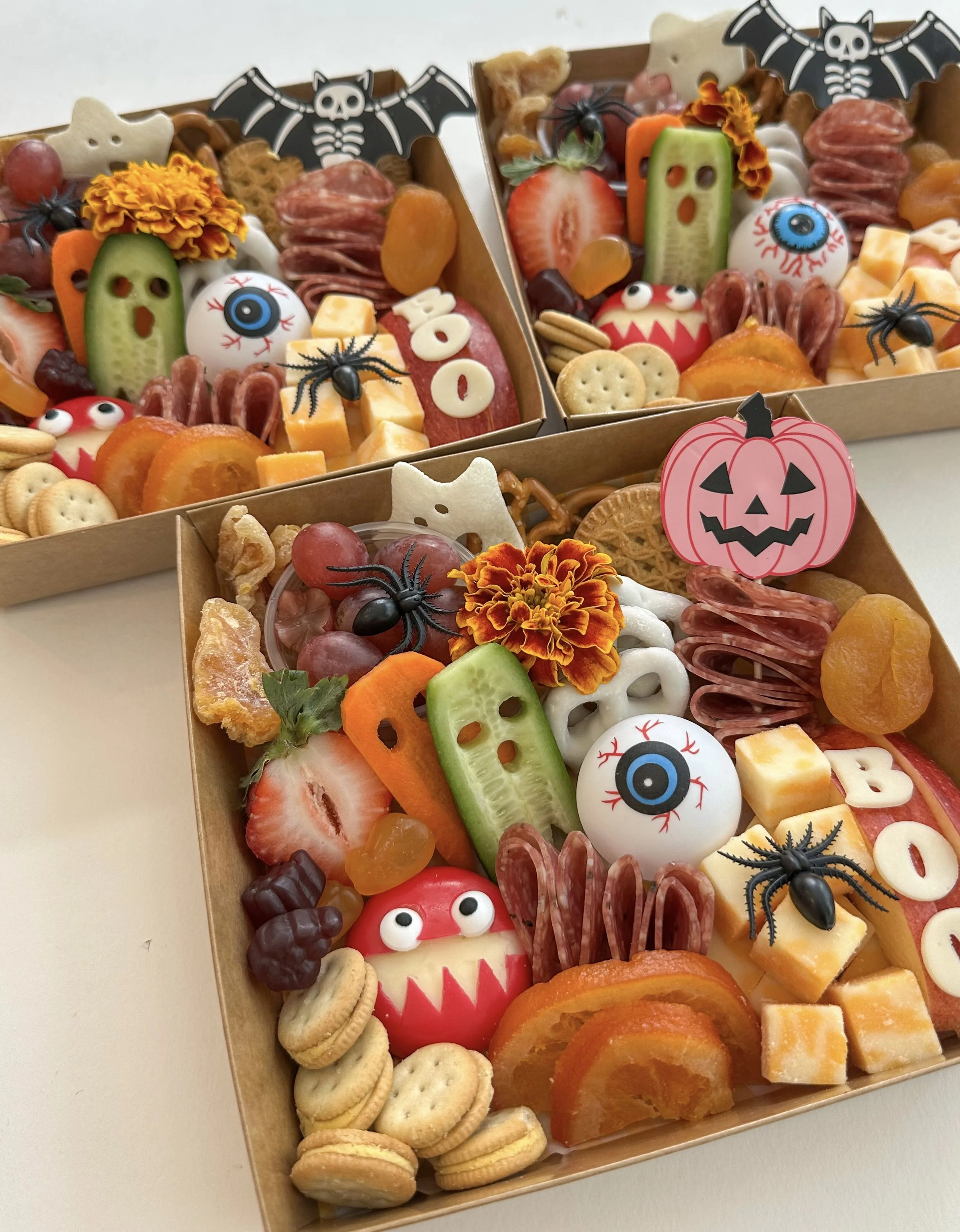 Three decorated Halloween-themed charcuterie boxes with candies, crackers, fruit, and themed decorations including googly eyes, spiders, ghosts, bats, and pumpkins.