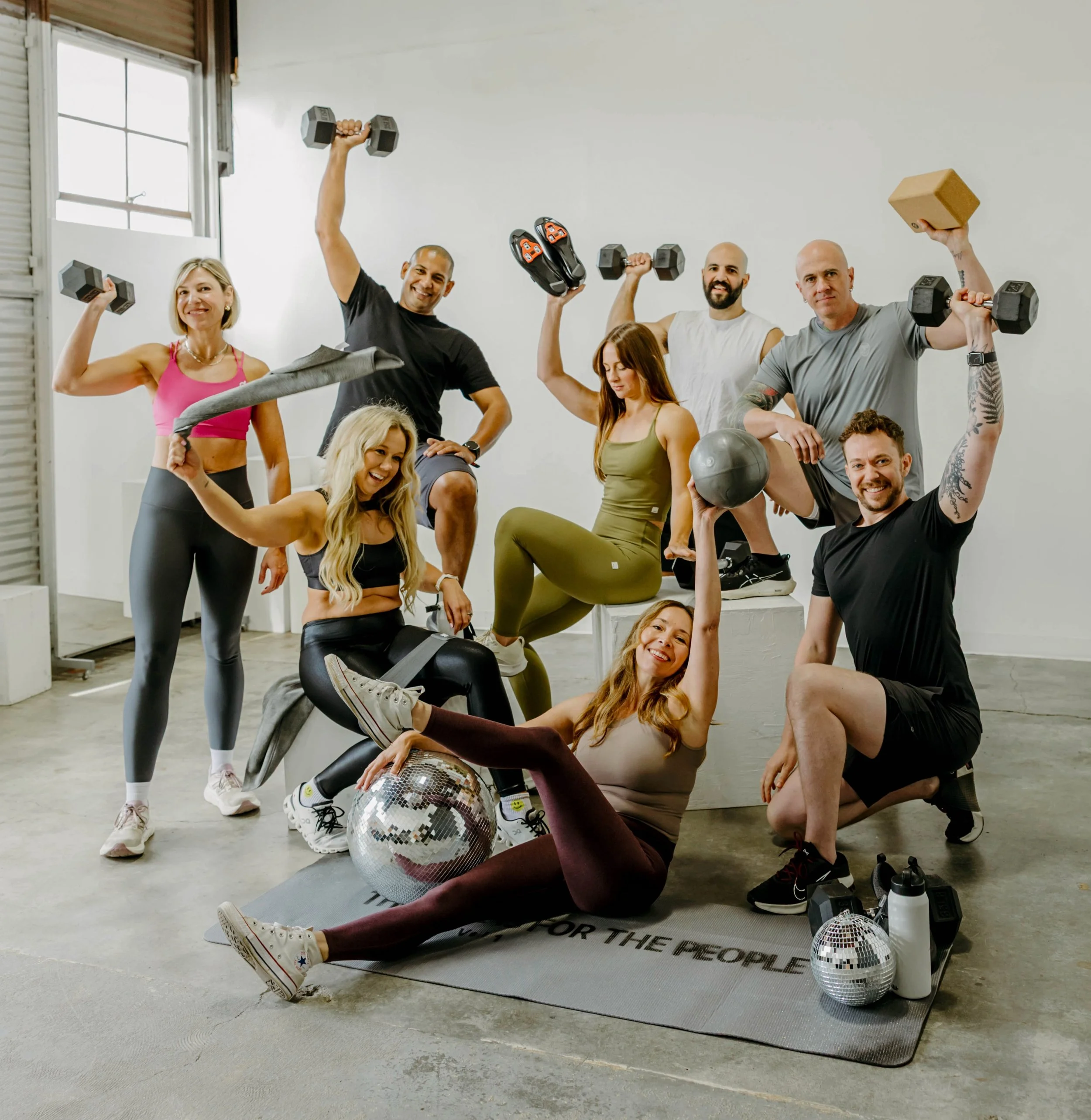 Group of people in workout gear with disco ball posing having fun with weights, yoga blocks, Pilates props, cycling shoes, and towels.