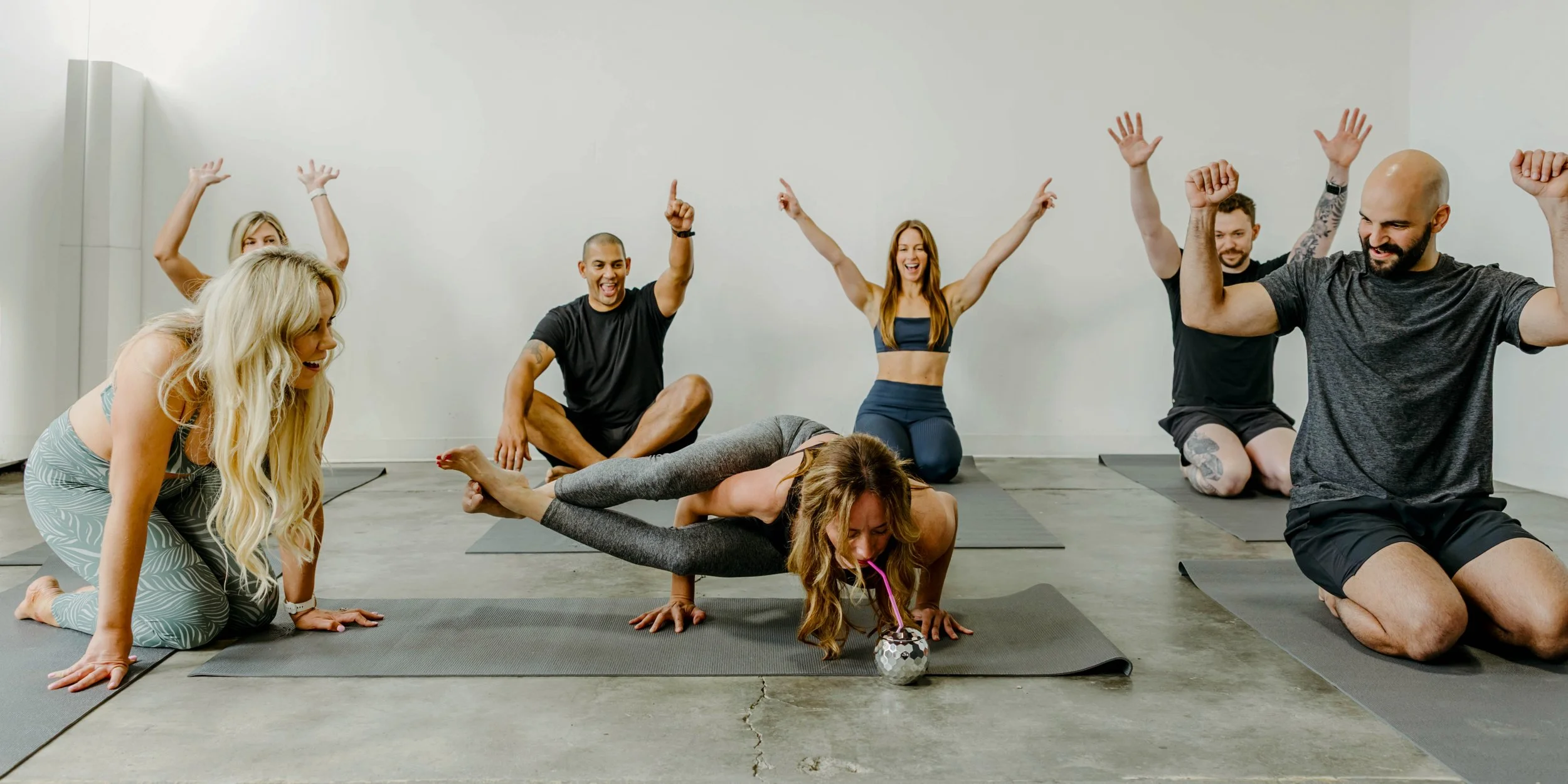 Woman doing a yoga pose drinking out of a disco ball and a group of people seated cheering her on.