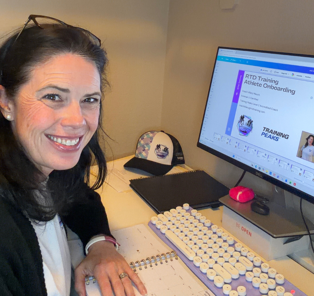 A woman with dark hair smiling at a desk, with a computer showing a presentation titled 'RTD Training Athlete Onboarding'. She is wearing glasses on her head, a white shirt, and a black jacket. On the desk are a keyboard, a notebook, a cap, and some papers.