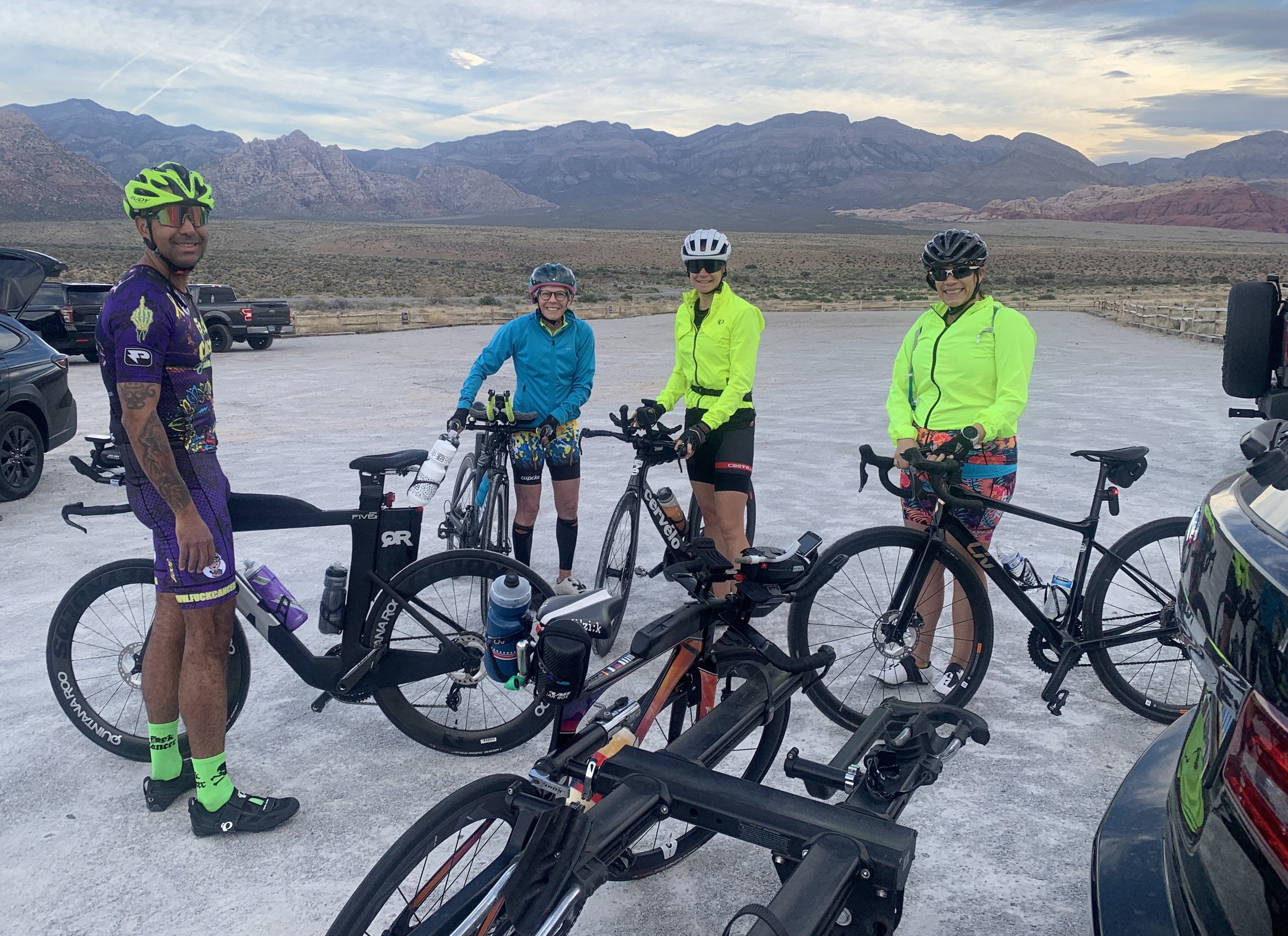 Four cyclists, two women and two men, stand with their bikes in a desert landscape with mountains in the background. They are dressed in colorful cycling gear, with helmets and sunglasses, smiling at the camera.