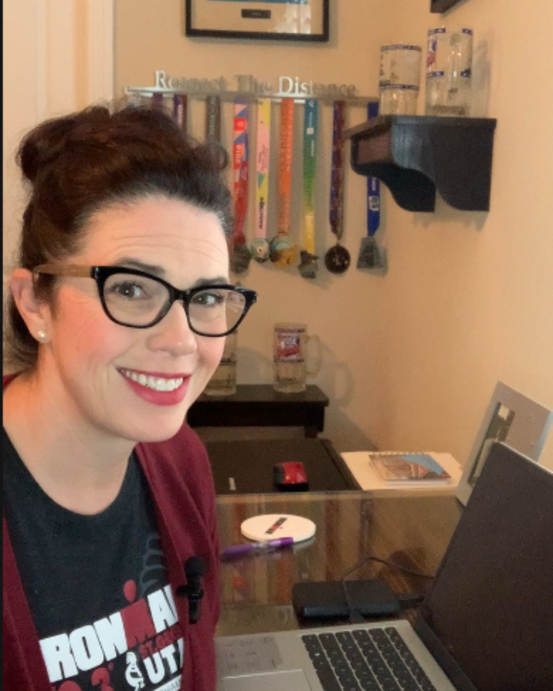 A woman with glasses and a dark top smiling in an office or home workspace with a laptop on a glass desk, water bottles on a black shelf, and a bulletin board with medals in the background.