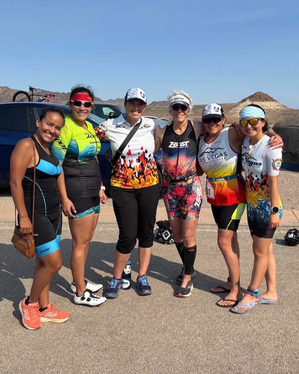 Group of six women in running or triathlon gear standing together outdoors in a desert landscape with mountains in the background, smiling at the camera.