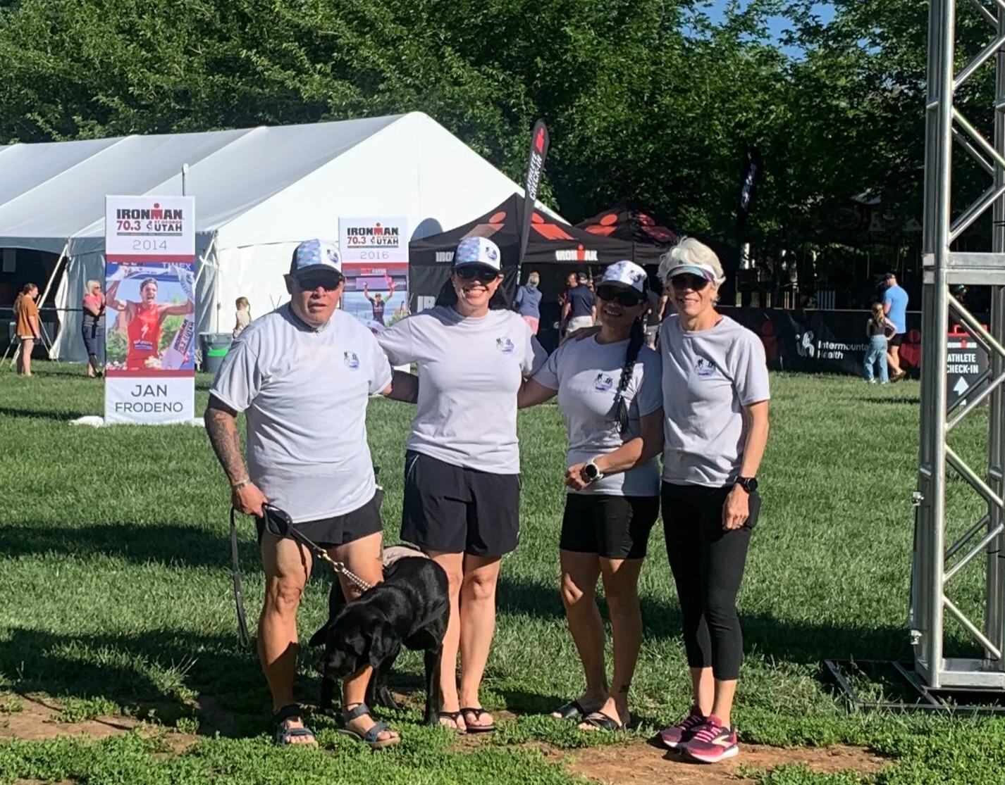 Four women standing outdoors on grass at a race event, with tents and banners in the background. One woman has a dog, and all are wearing athletic clothing and sunglasses.
