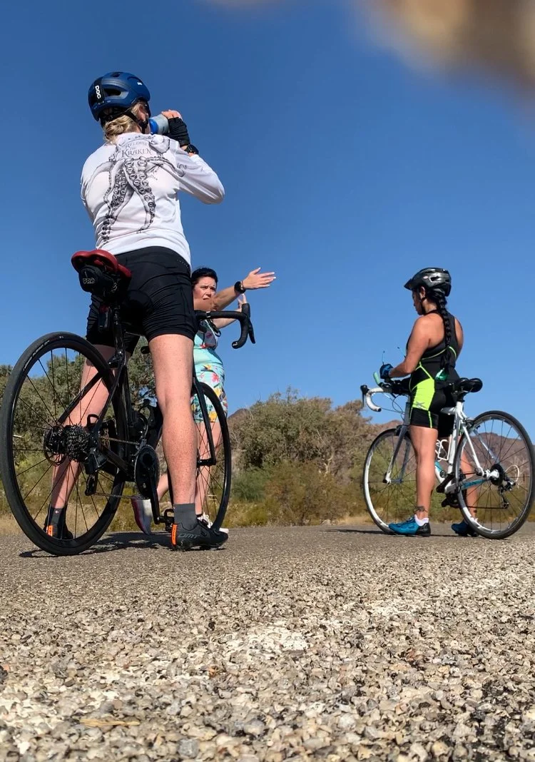 Three cyclists, two men and one woman, standing on a paved road outdoors, with trees and bushes in the background, one woman gesturing while talking to a man on a bicycle, all wearing helmets and athletic apparel.