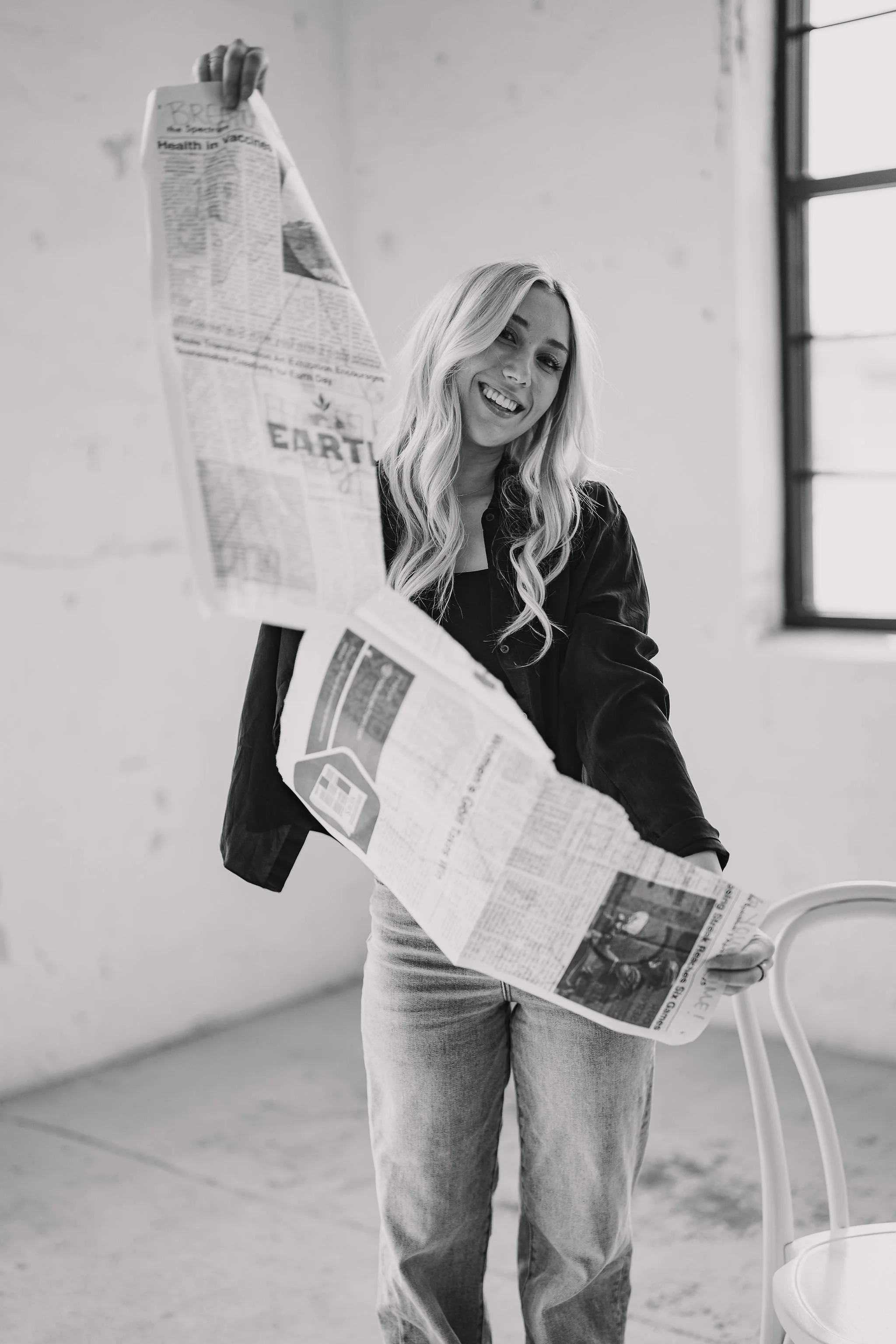 A smiling young real estate agent with long wavy hair wearing a dark shirt and jeans, holding a crumpled newspaper in an industrial-style room with a white wall, large window, and a white chair.