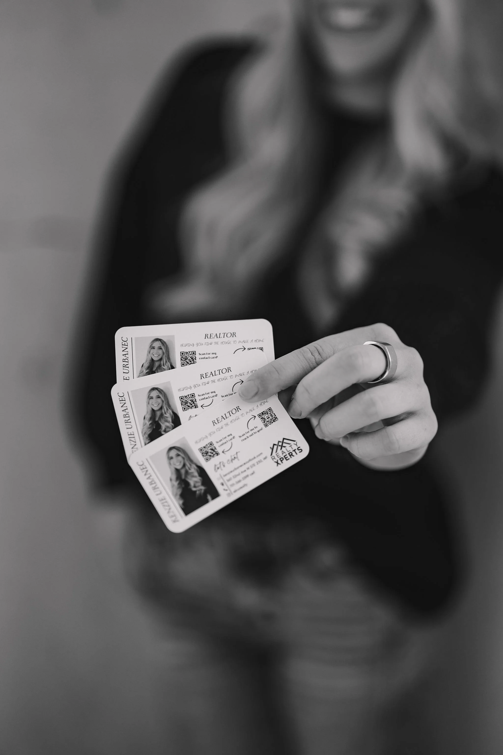 A realtor holding three real estate agent business cards, each featuring her photo, QR code, and contact information, in black and white.