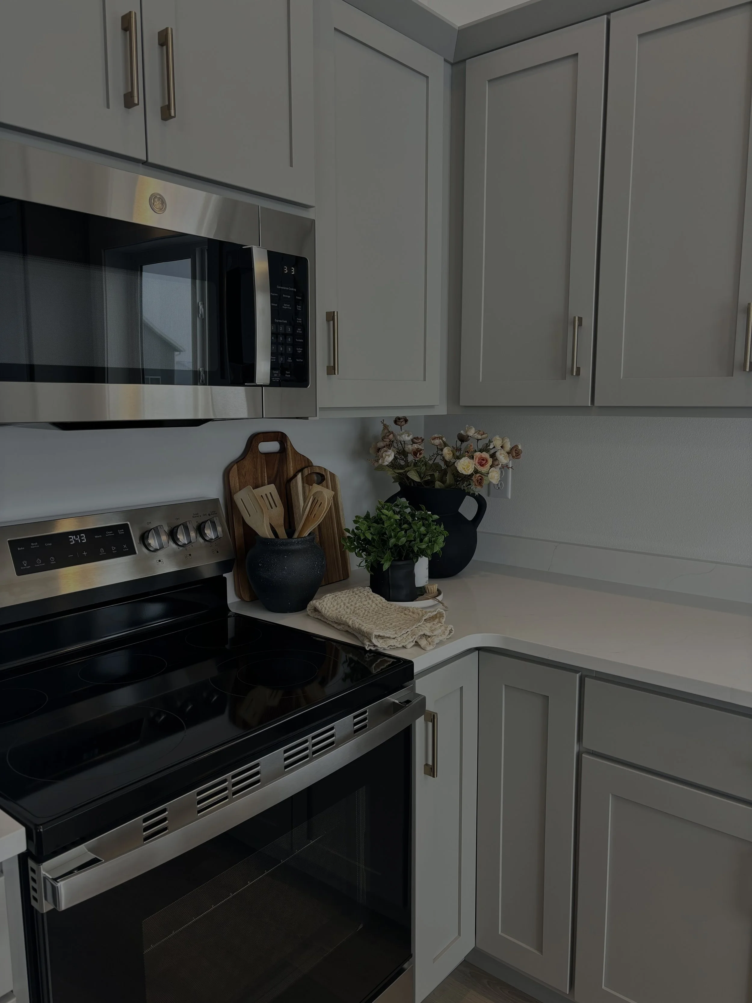 Kitchen corner with grey cabinets, stainless steel microwave and oven, black and wood utensils on the counter, and decorative flowers in black vases. new construction home