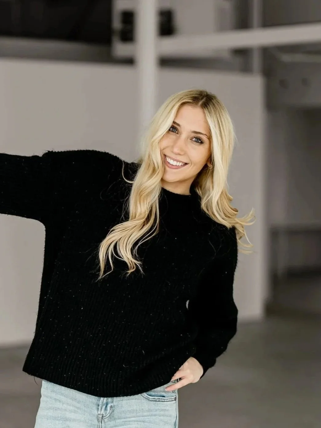 A young realtor with long blonde hair, smiling, wearing a black sweater and light blue jeans, indoors with a modern, minimalist background.