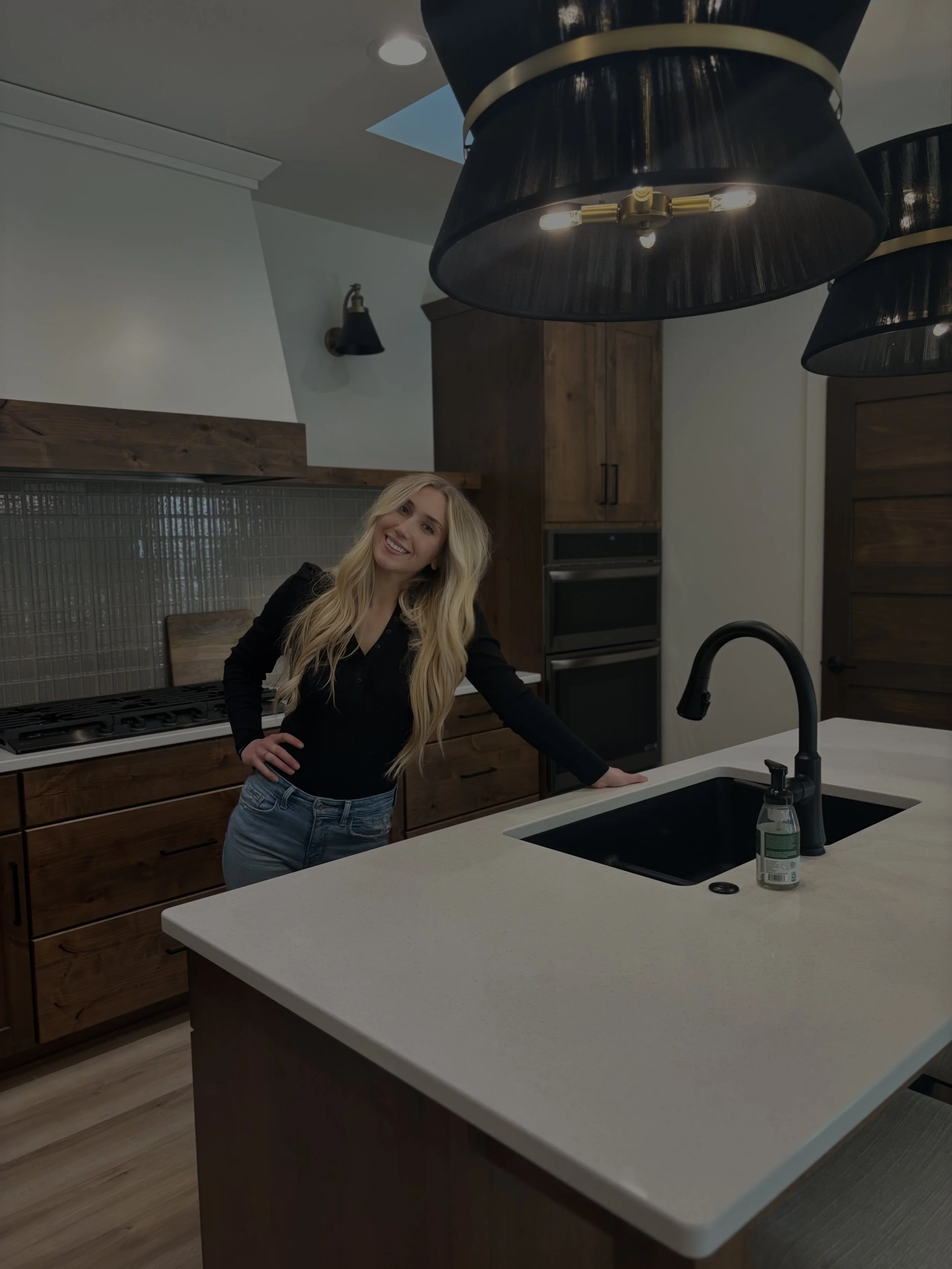 A realtor with long blonde hair smiling while standing in a modern kitchen by a white countertop with a black sink and black faucet, surrounded by wooden cabinets, with black ceiling lights overhead.