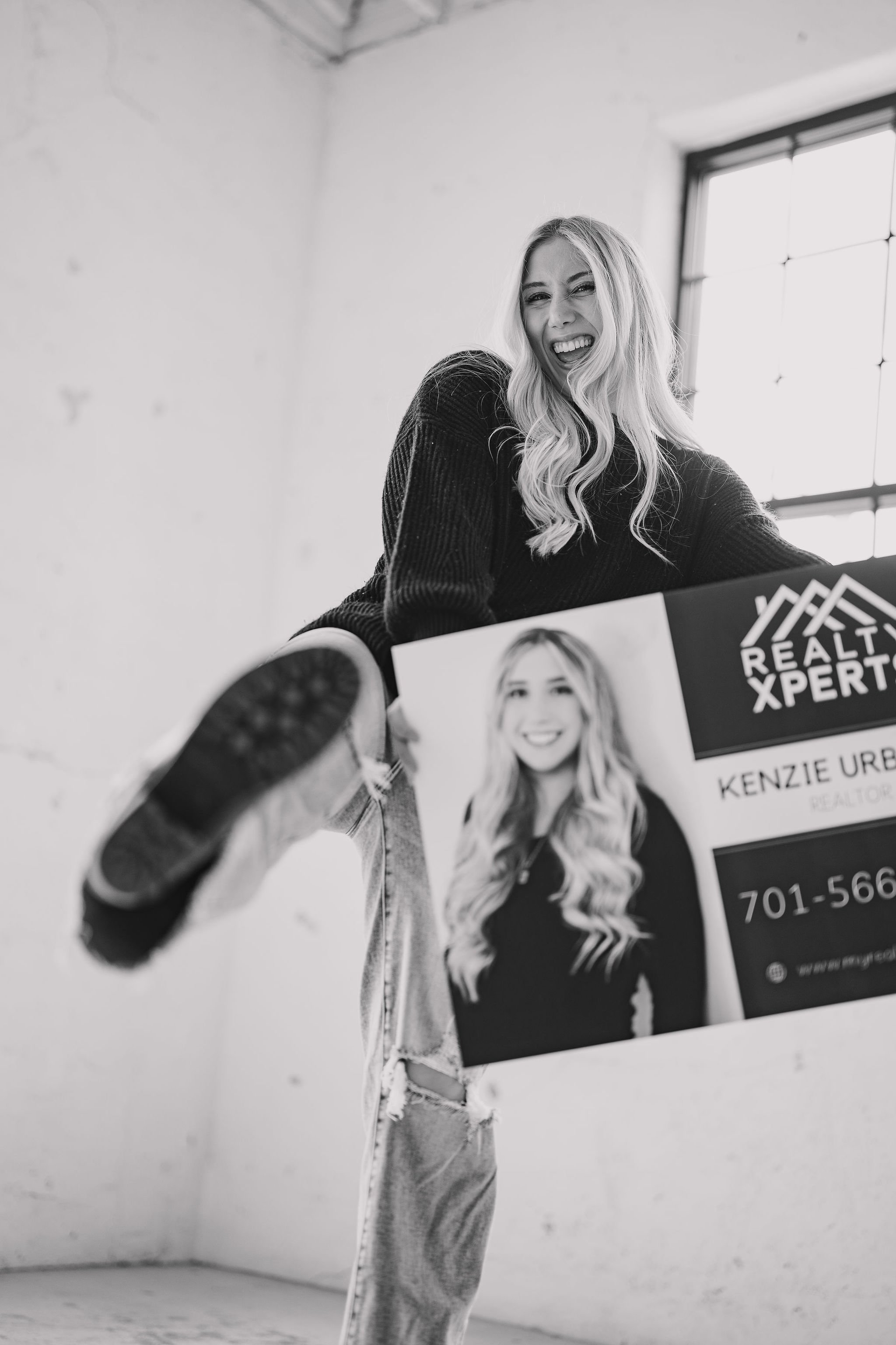 A woman with real estate agent wavy hair smiling and holding a real estate sign with her picture and contact information, indoors near a large window.