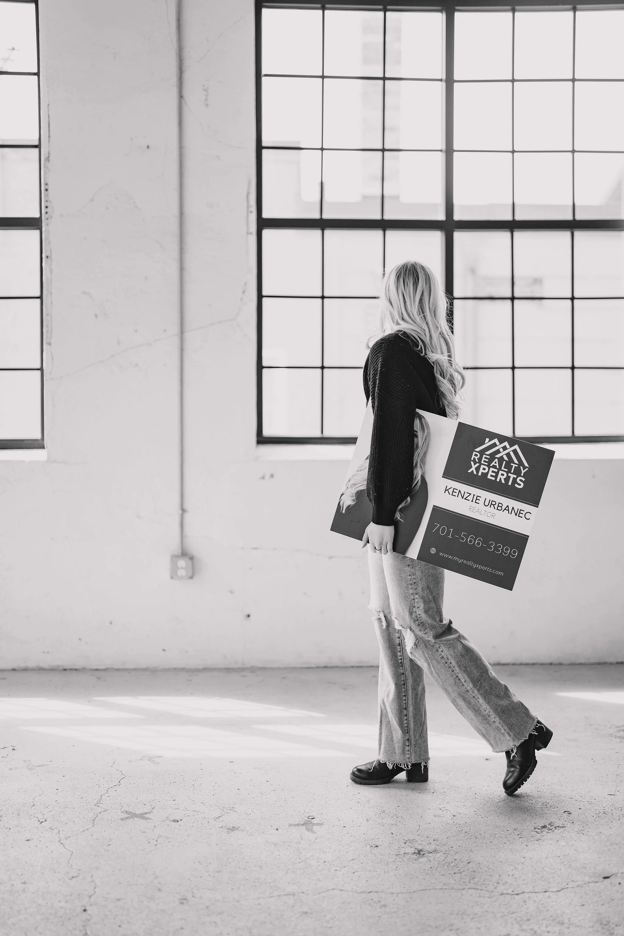 A real estate agent with long hair holding a real estate sign in an industrial-style room with large windows.