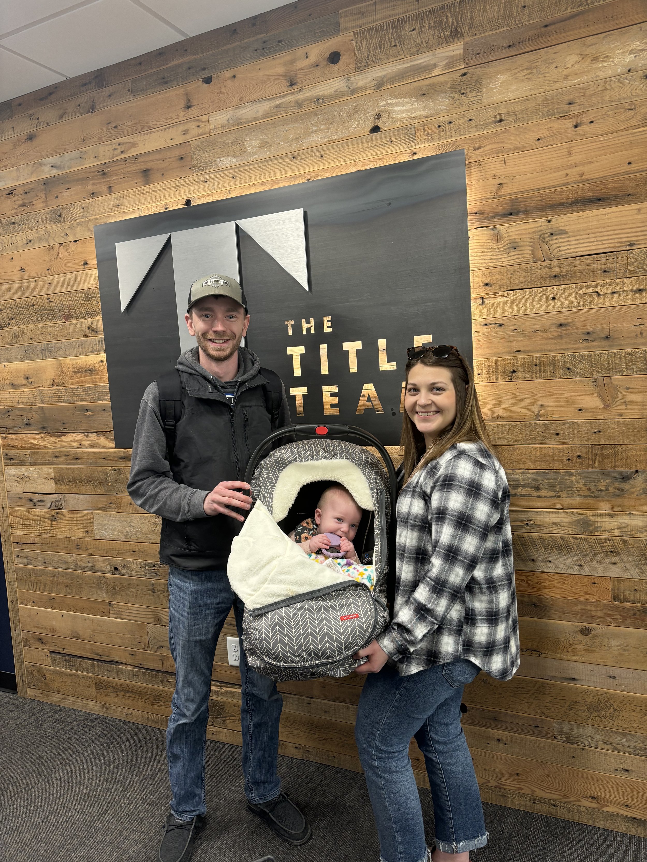 A smiling man, woman, and a baby in a stroller are standing in front of a wall with a wooden panel background and a sign that says 'The Title Team'. real estate closing