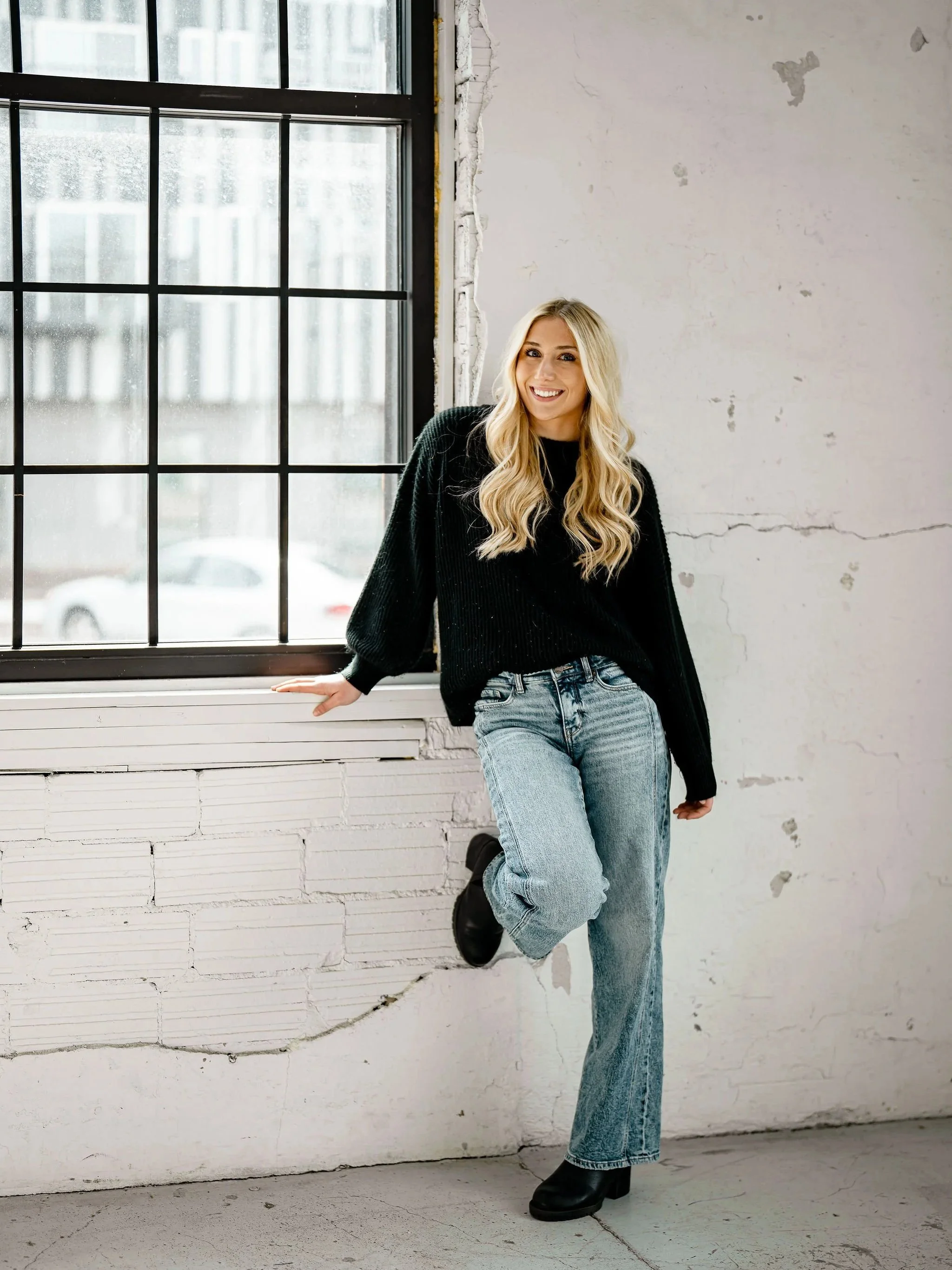 A young realtor with long blonde hair and a bright smile, wearing a black sweater, light blue jeans, and black boots, leaning against a white brick wall with a large window in the background.