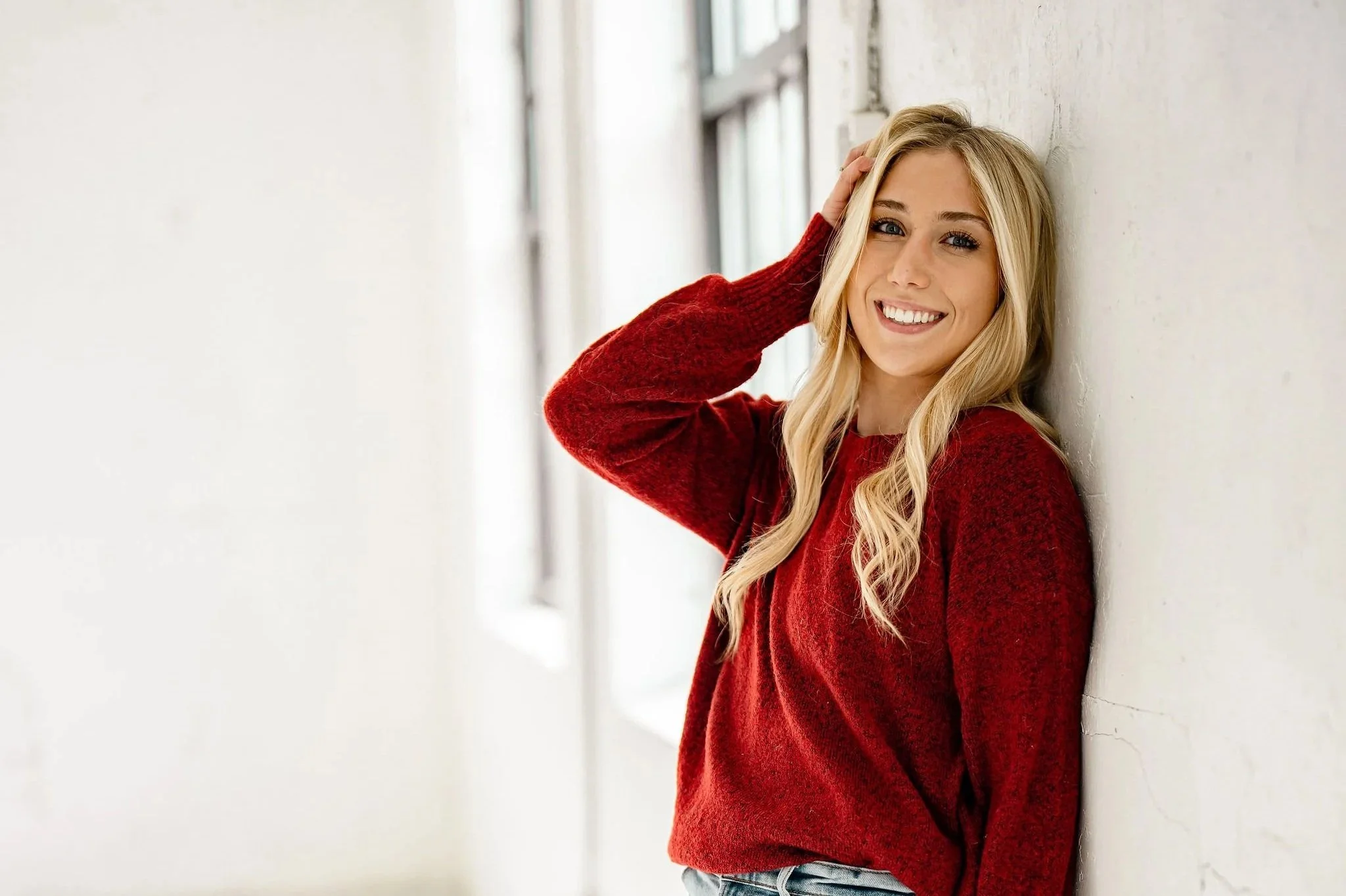A young realtor with long blonde hair, wearing a red sweater, smiling, leaning against a white wall in a hallway.