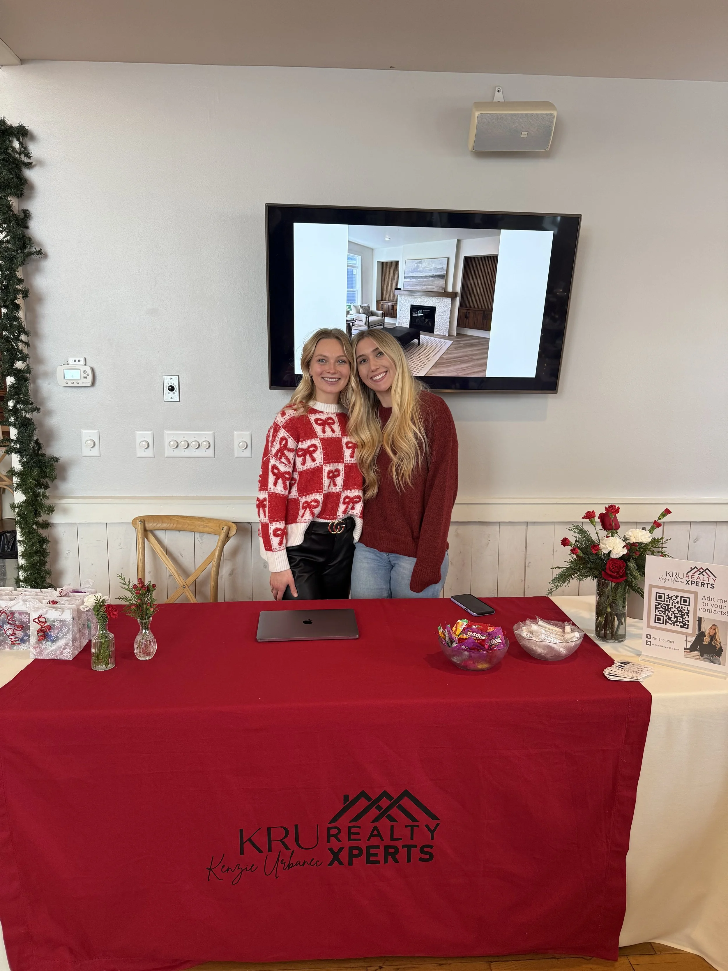 Two smiling women standing behind a table with a red tablecloth that has the logo 'KRU REALTY XPERTS' on it. They are in a room decorated for the holidays, with presents, flowers, and marketing materials on the table. A large TV screen behind them displays an interior room with a fireplace.
