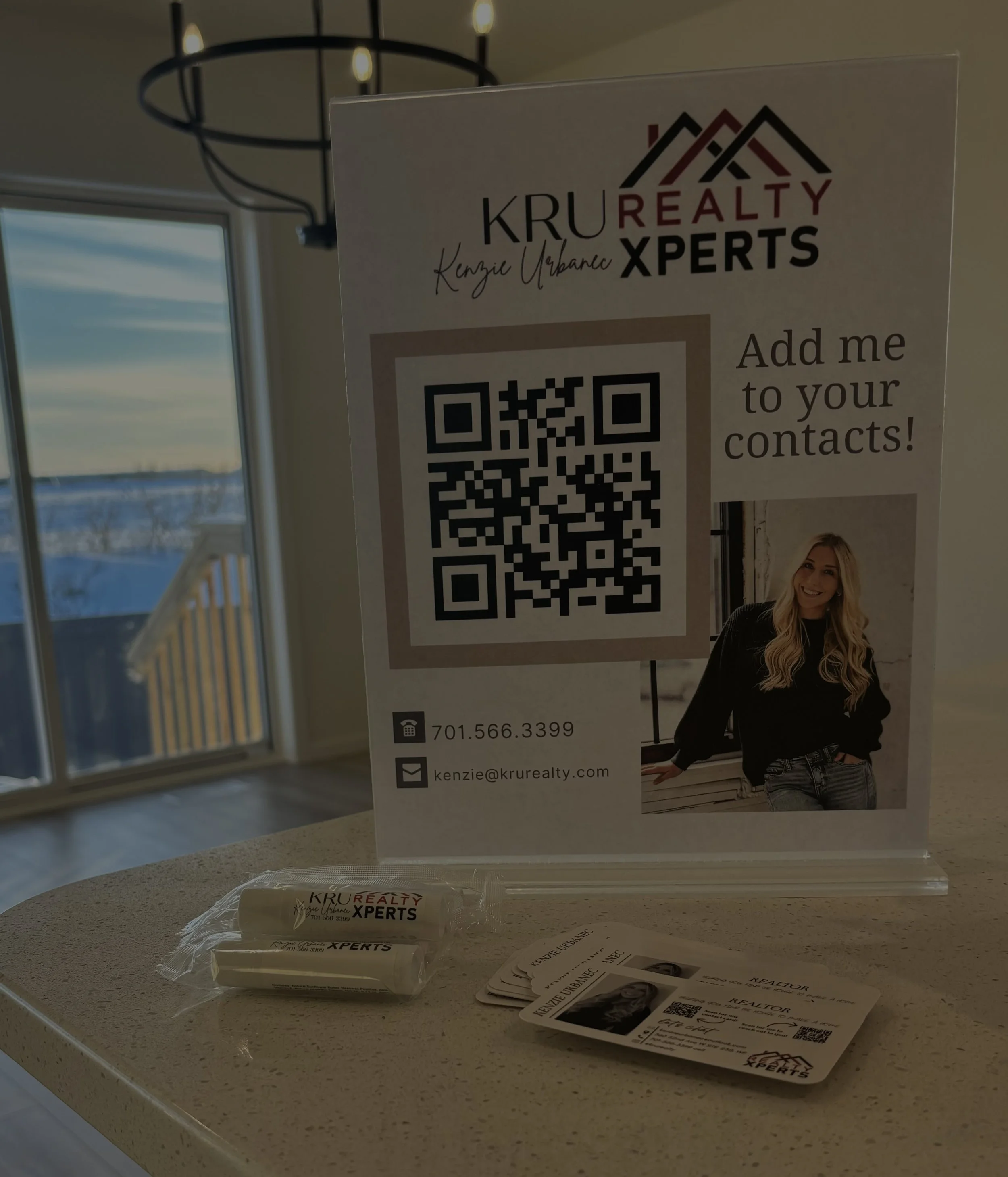 A real estate promotional display at open house featuring a QR code, business contact information, a woman with blonde hair smiling, and real estate business cards and branded items on a countertop in a well-lit room with a window and view outside.