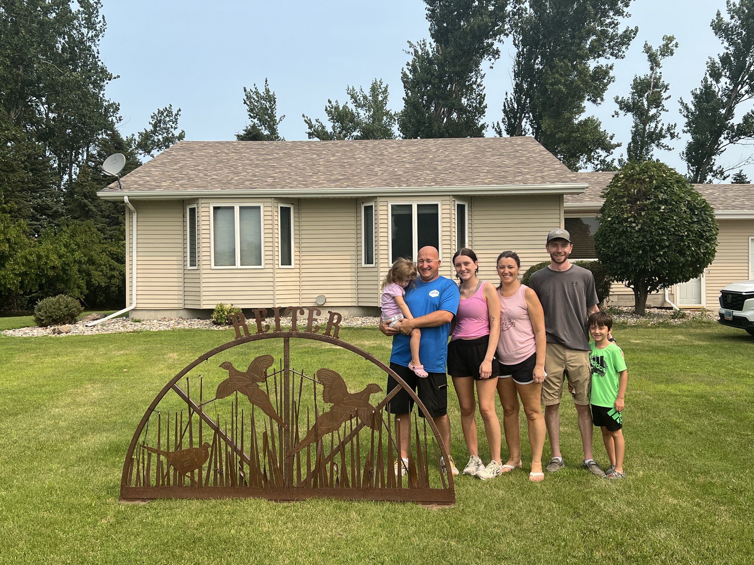 family buying a new house. A family of five standing on a lawn in front of a house, with a decorative metal sign that says "Reiter" featuring flying birds and a rabbit.