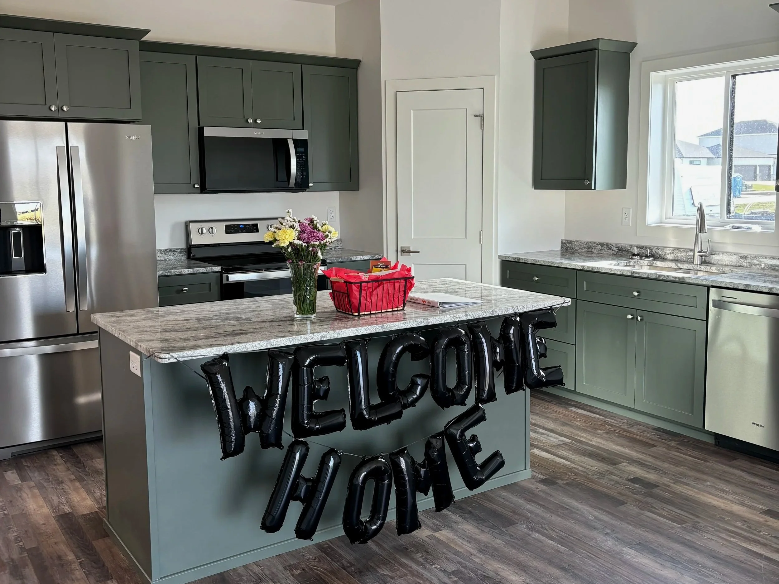 Kitchen with green cabinets, stainless steel refrigerator, microwave, and stove. An island with a bouquet of flowers and black balloons spelling 'WELCOME HOME' is in the center. A window with outside houses is above the sink. realestate closing gift