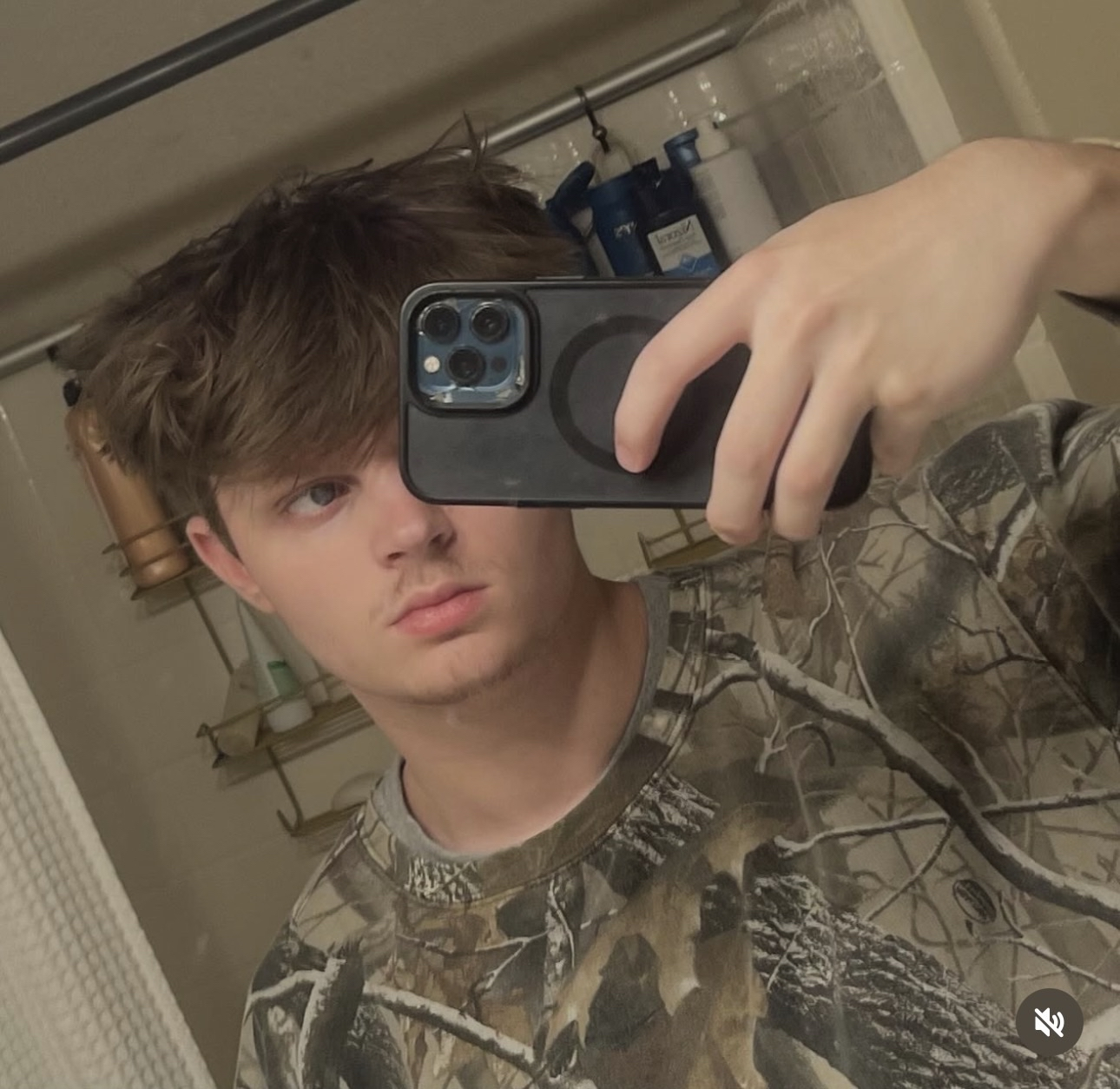 Violxt Flowers taking a mirror selfie with a smartphone in a bathroom. He has light brown, slightly messy hair, and is wearing a camouflage shirt.