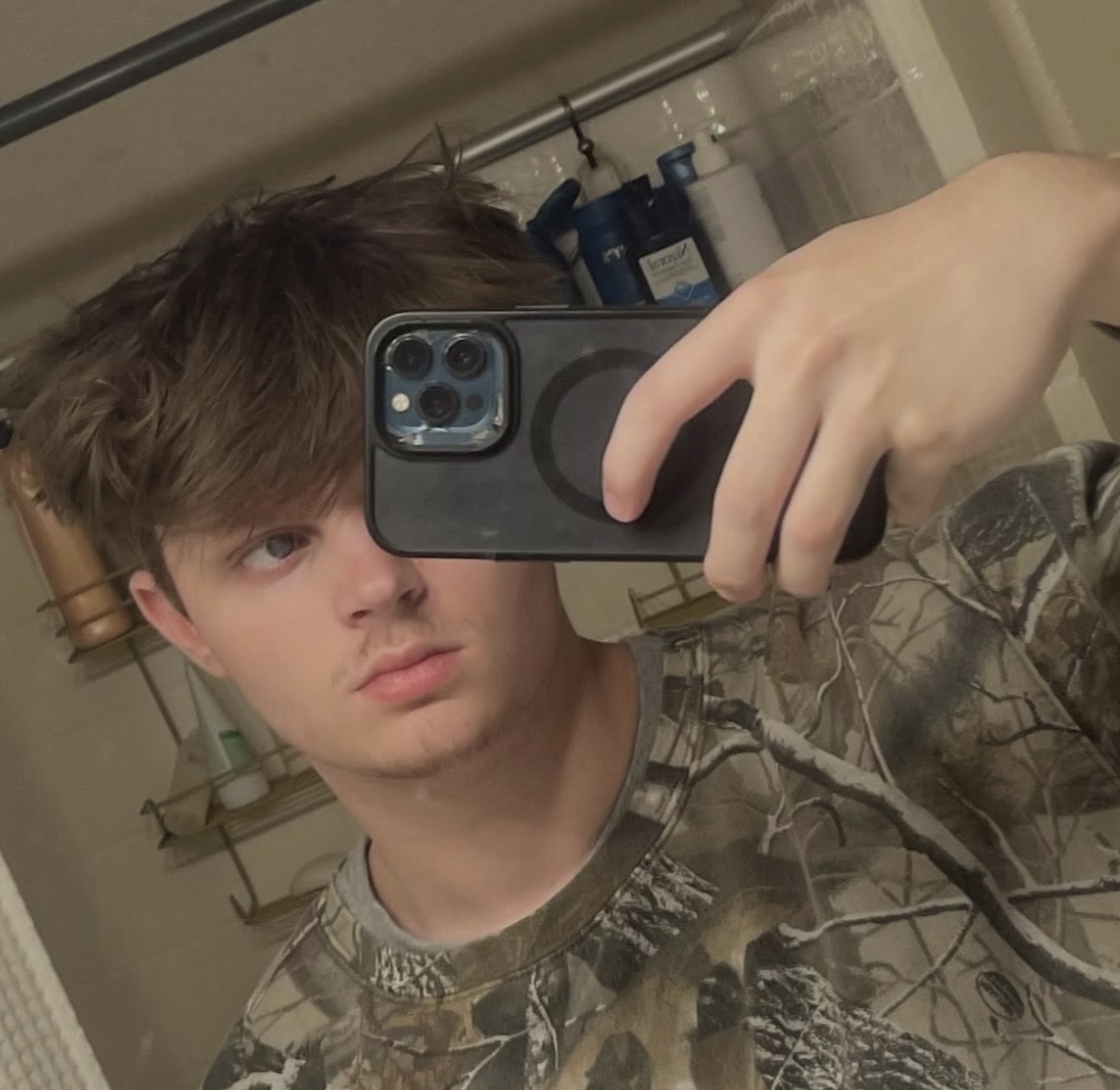 Violxt Flowers taking a mirror selfie with a smartphone in a bathroom. He has light brown, slightly messy hair, and is wearing a camouflage shirt.