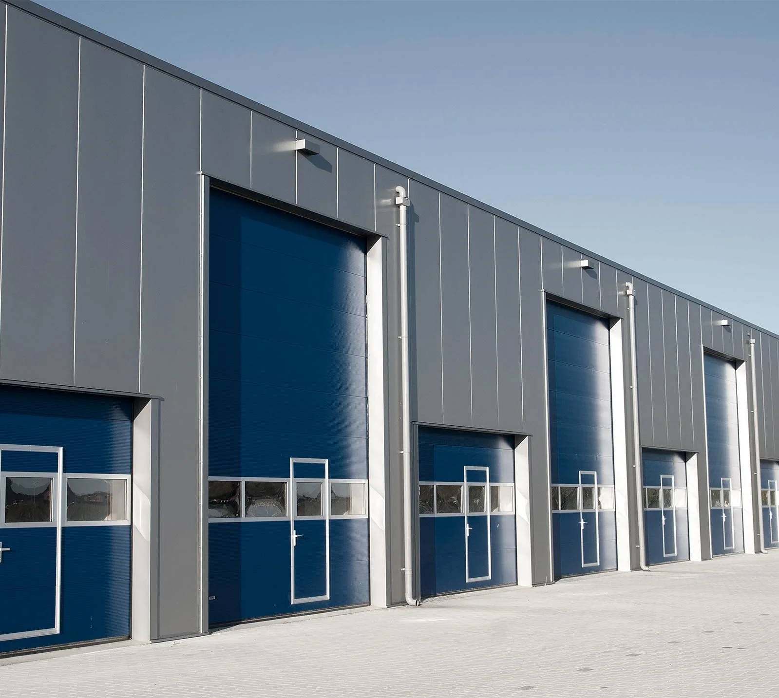 A row of industrial warehouses with large blue garage doors and gray metal siding under a clear blue sky.