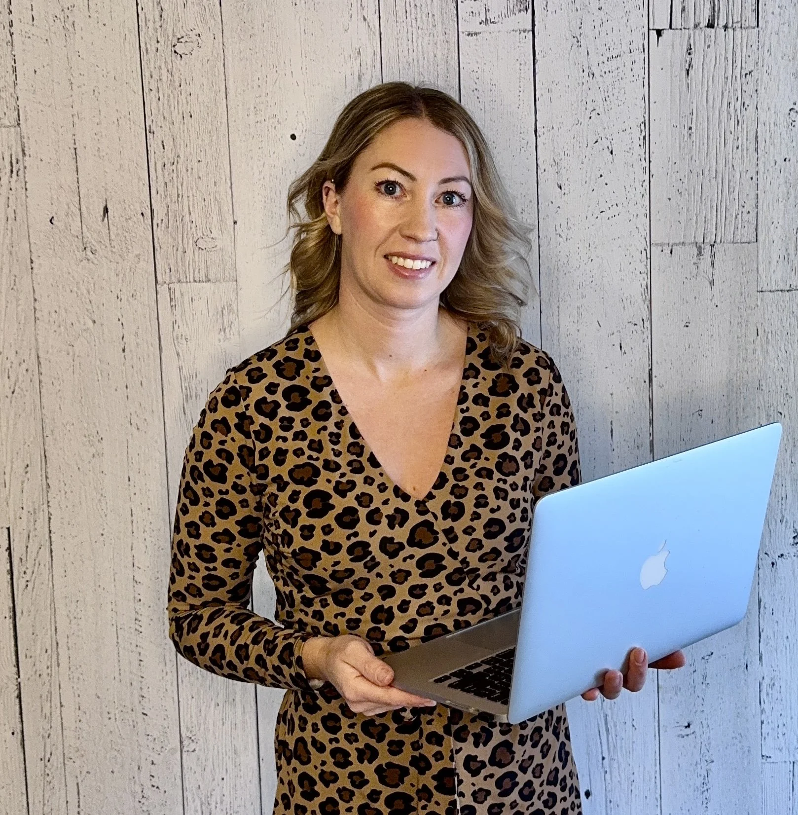 A woman with blonde, wavy hair wearing a leopard print dress, holding a silver MacBook, standing in front of a light wooden wall.