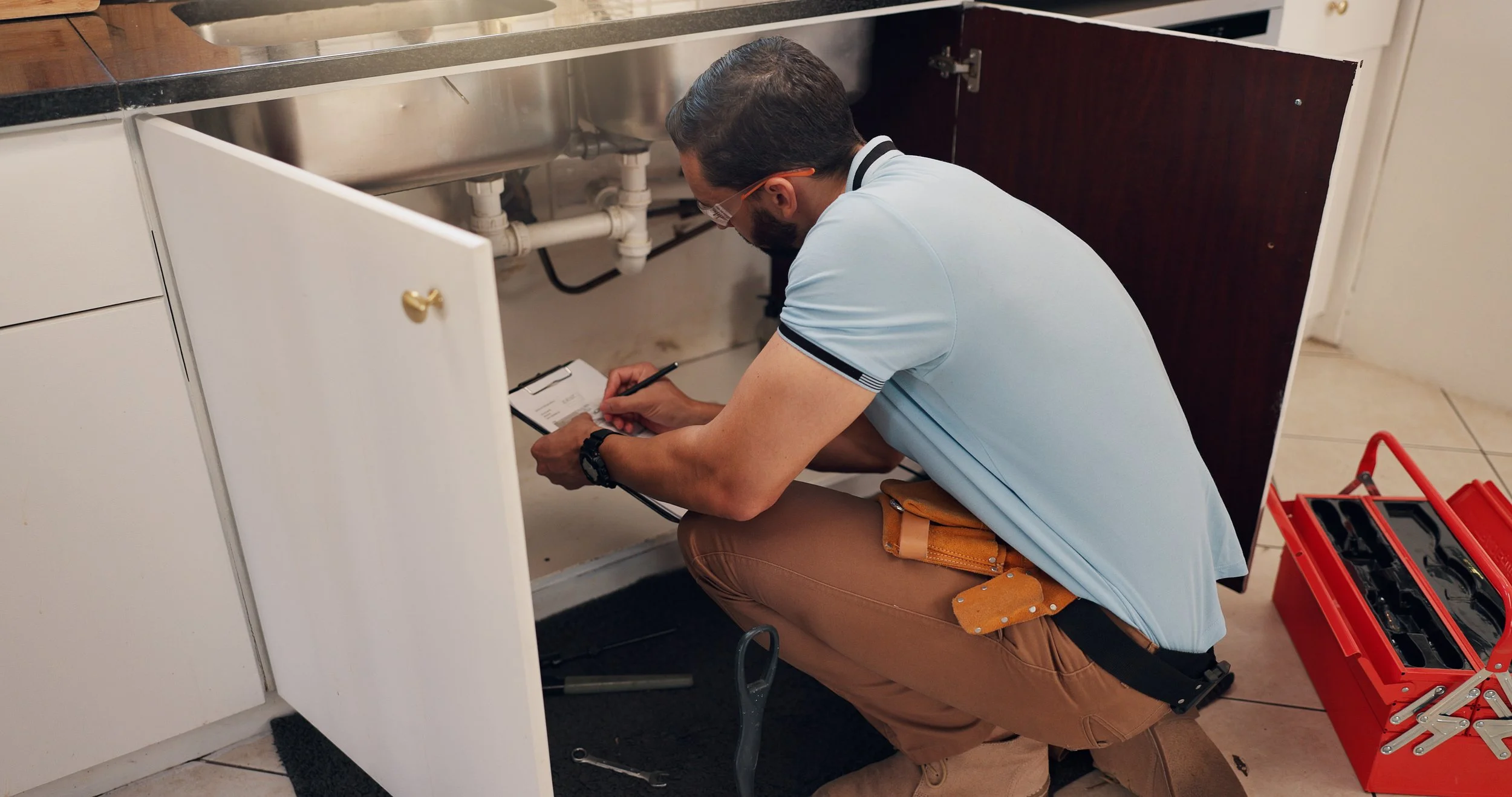 Man with a clipboard inspecting pipes under a sink