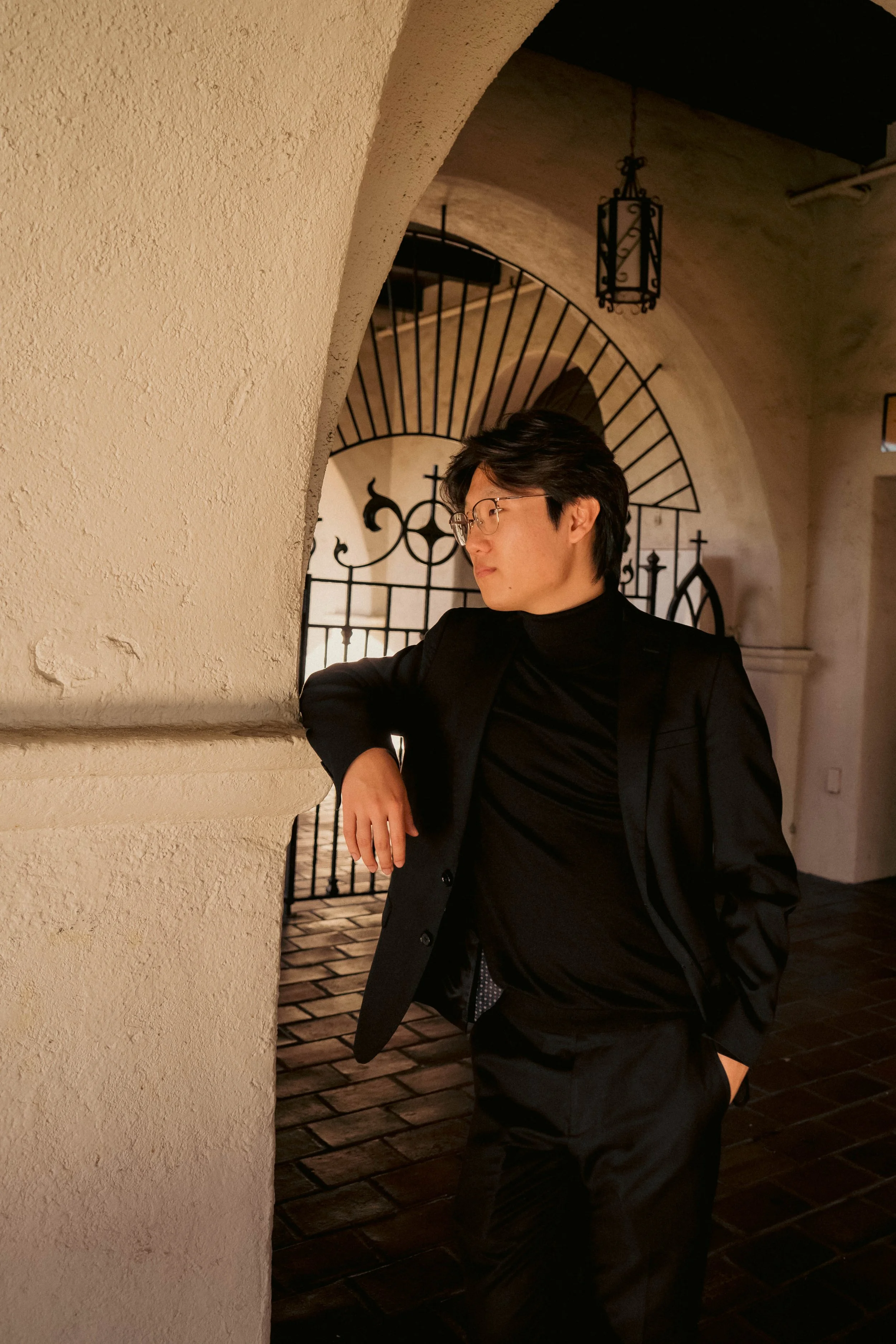 A young man in a black suit and turtleneck, wearing glasses, leaning against a white textured wall in an elegant, dimly lit interior with gothic-style black iron gate and hanging lanterns.