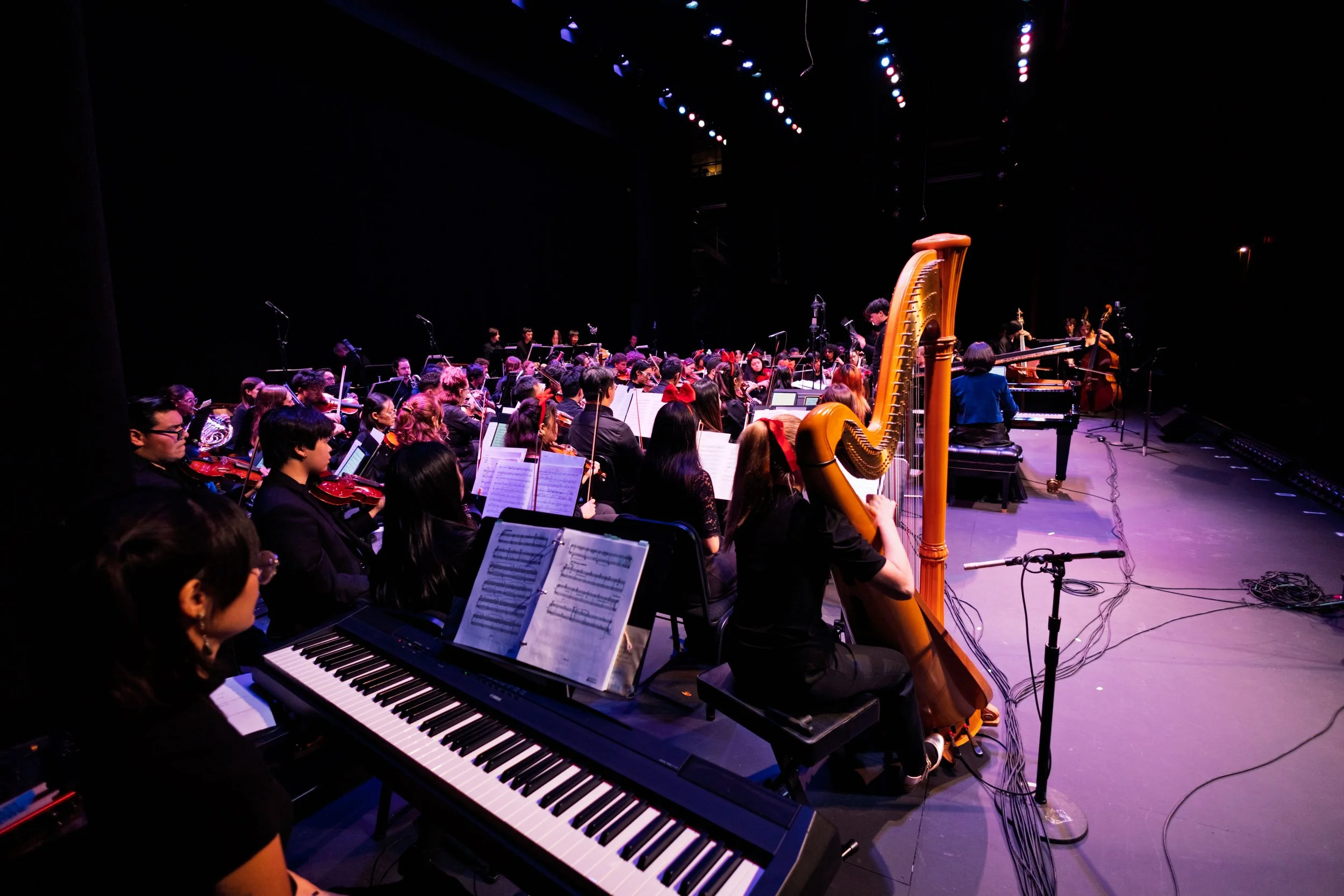 San Gabriel Symphony Orchestra on stage during a performance, with musicians playing string, wind, and keyboard instruments, on a dark lit stage with a black background.