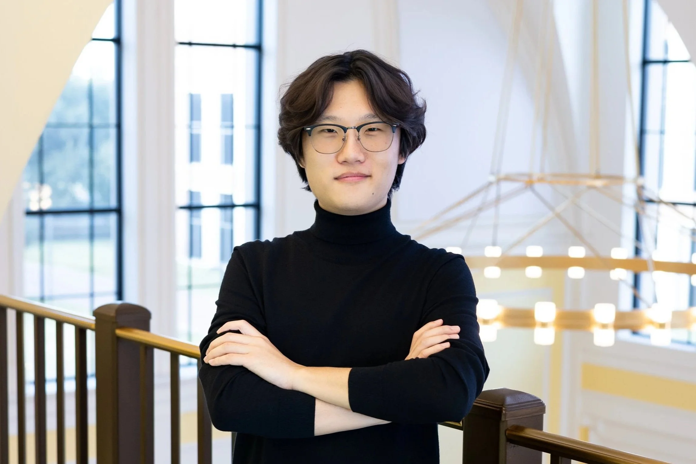 William Jae with glasses wearing a black turtleneck, standing indoors with arms crossed, in front of large windows and a modern chandelier at the Shepherd School of Music.