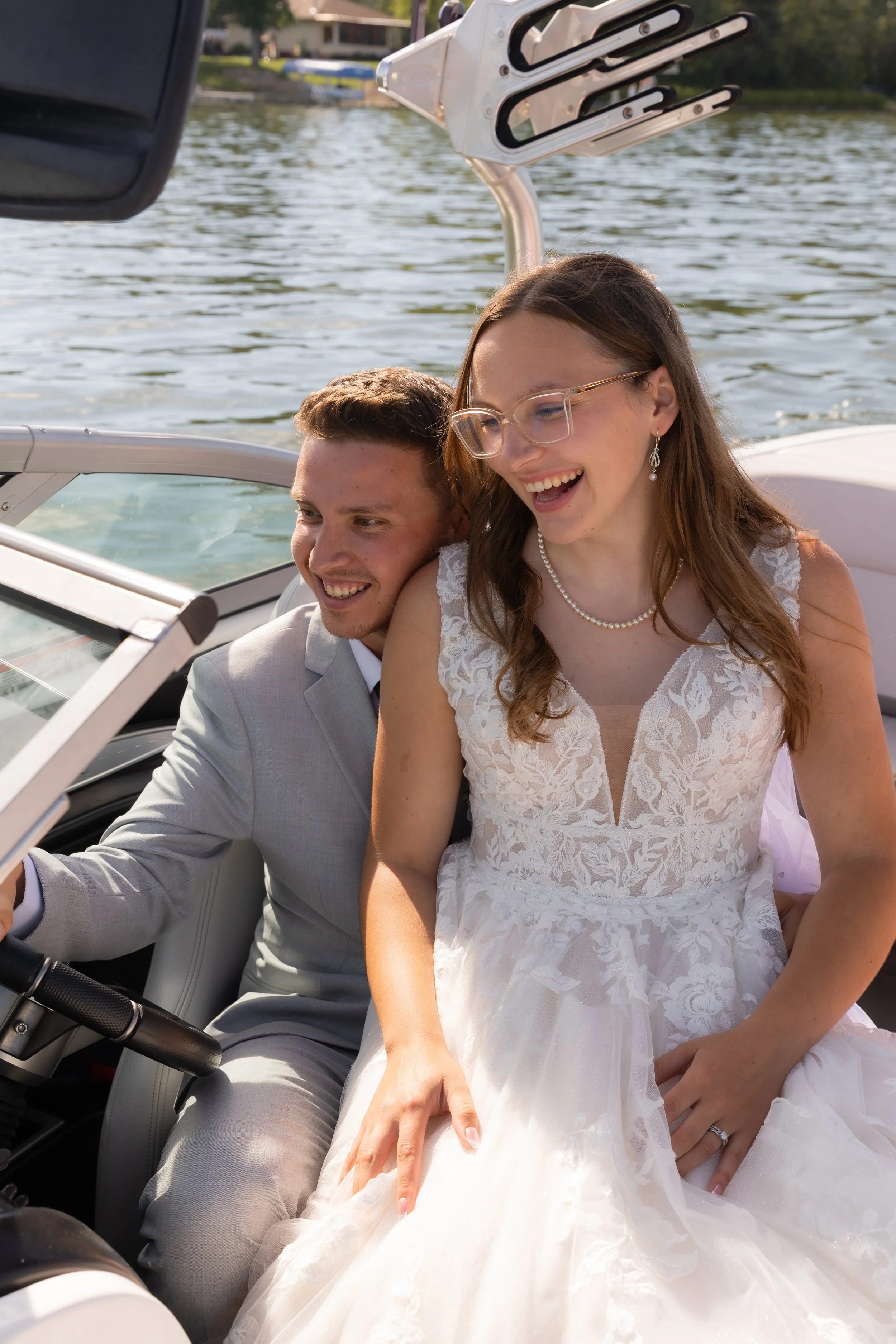 A newlywed couple on a boat, smiling and enjoying their day by the water. The bride wears a white lace wedding dress and pearl jewelry, while the groom wears a light gray suit. The background shows a river with a house and green trees.
