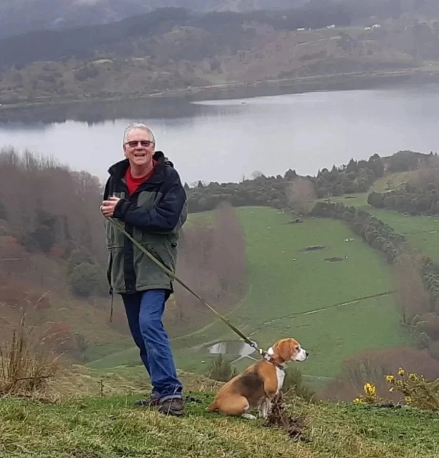 John Henton, stands on a grassy hill, smiling, holding a leash attached his beagle dog, "Giggle" with a lake and rolling hills in the background.