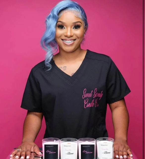 Smiling woman with blue wavy hair wearing a black medical scrub top with pink embroidery, standing behind four glass containers filled with different colored liquids, against a pink background.