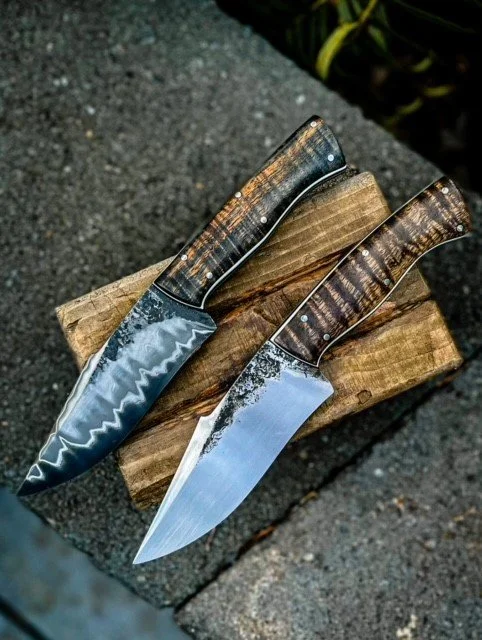 Two knives with wooden handles and decorative blades resting on a piece of wood.