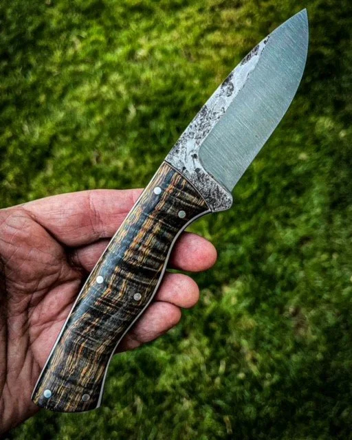 Hand holding a custom knife with a wooden handle and a textured blade, outdoors on grass.
