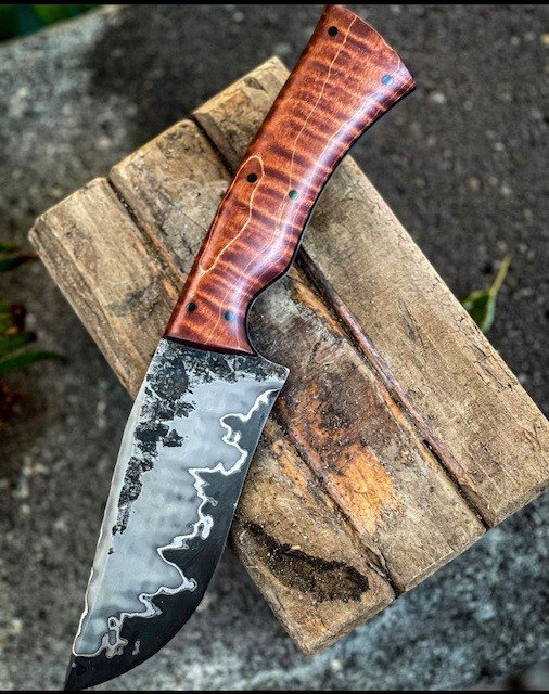 A custom-made hunting knife with a Damascus steel blade and a wooden handle, resting on a weathered piece of wood outdoors.