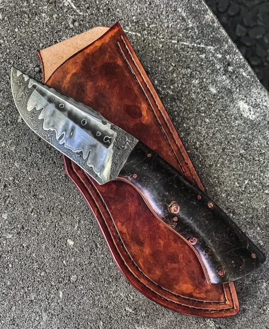 A large knife with a patterned, wavy edge and a dark, polished handle resting on a brown leather sheath on a stone surface.