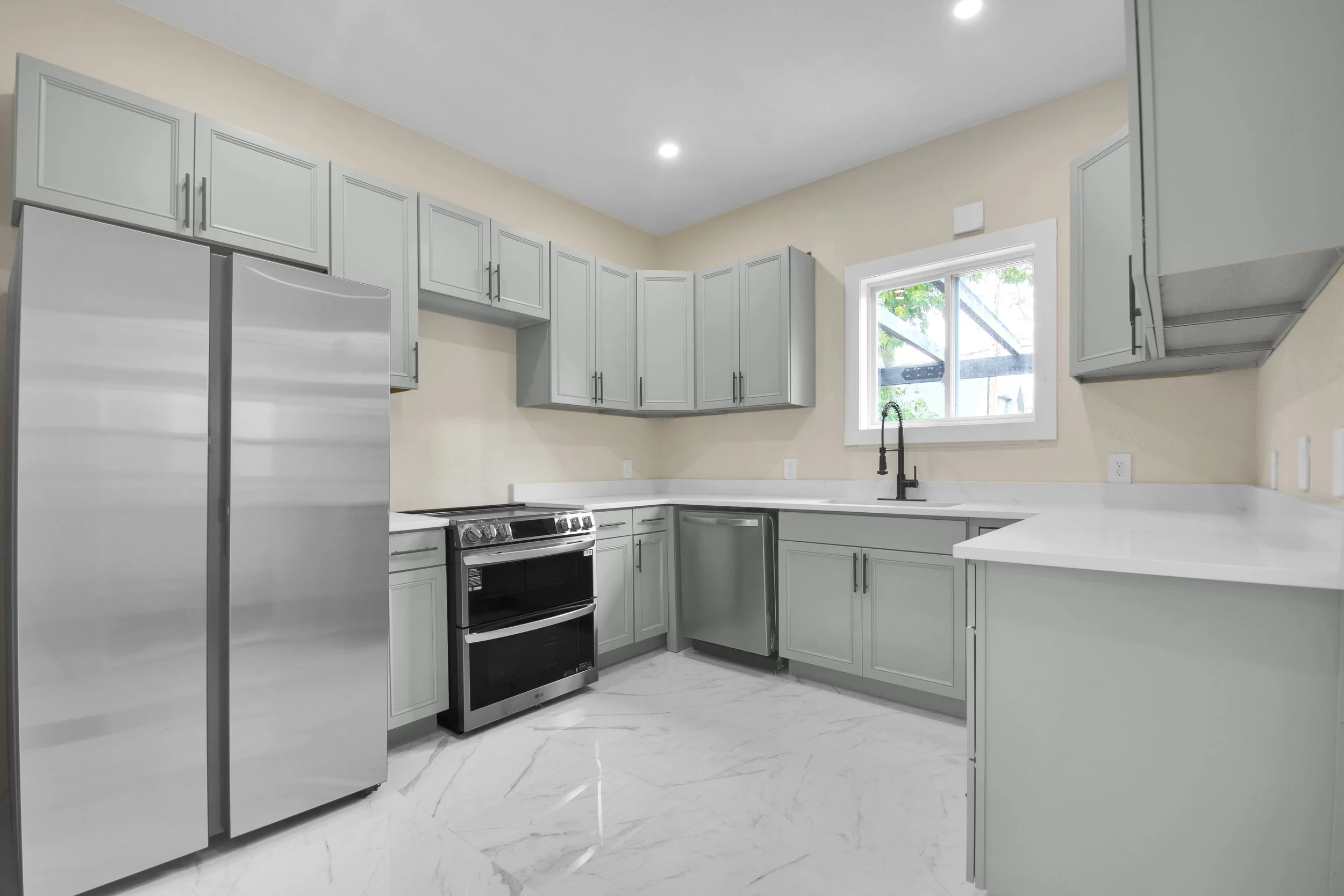Modern kitchen with light gray cabinets, stainless steel appliances including a refrigerator and stove, black sink faucet, white marble countertops, and a window overlooking greenery.